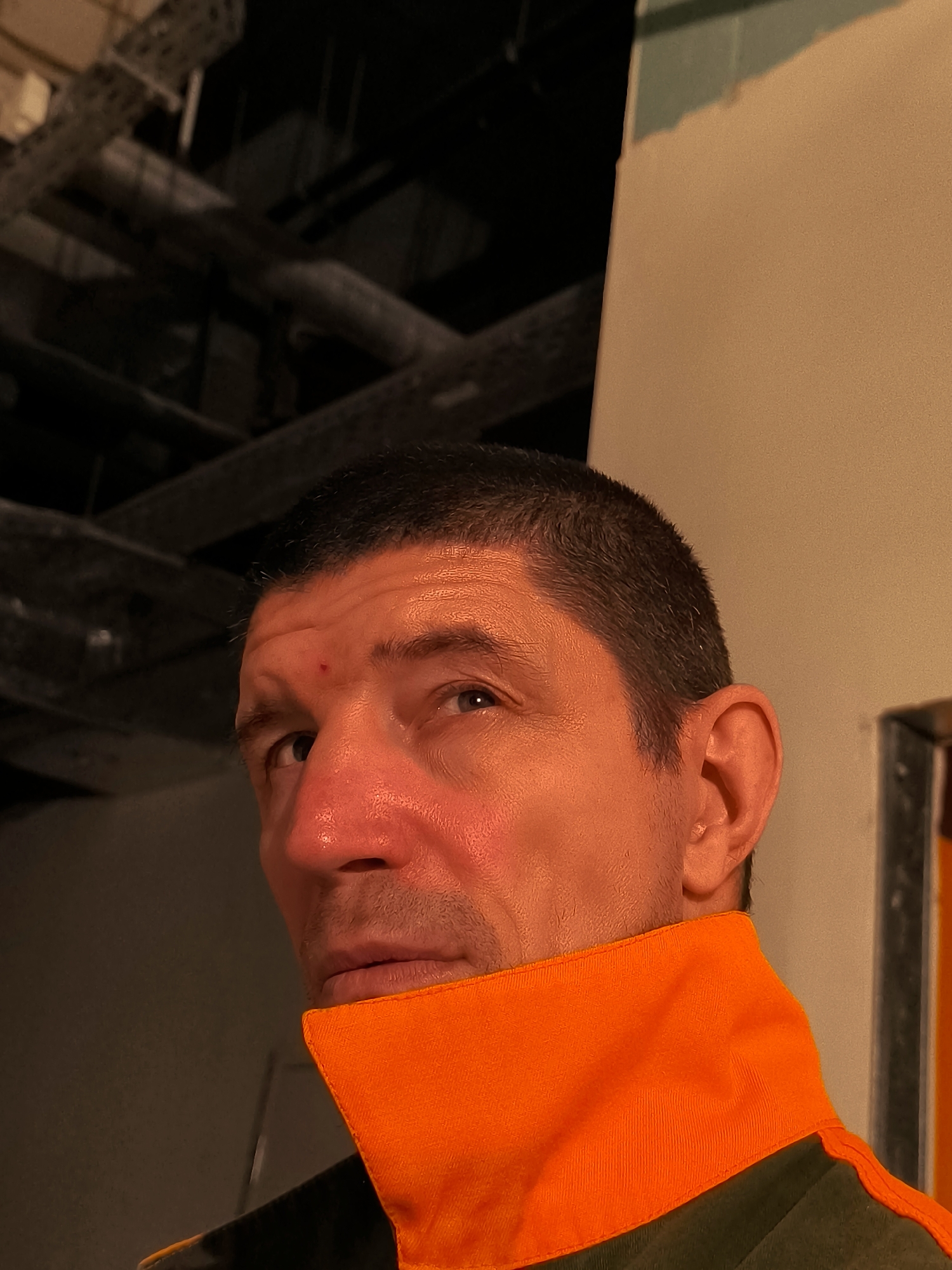 A close-up, eye-level shot captures the upper torso and face of a middle-aged man. He has short, dark hair and fair skin, with a small red mark on his forehead. His gaze is directed upwards and slightly to the left, towards a dimly lit area that appears to be an industrial or construction site, suggested by metallic conduits and beams visible in the upper left portion of the frame. The man is wearing a dark green garment with a prominent, bright orange collar. The lighting is warm and casts strong shadows, particularly on the right side of his face, highlighting the contours of his nose and cheekbone. The background transitions from the dark, industrial setting to a lighter, painted wall on the right. There is no clear indication of his location within the city of Qarağandı city administration, Kazakhstan, as the visual cues are generic to construction or industrial environments.