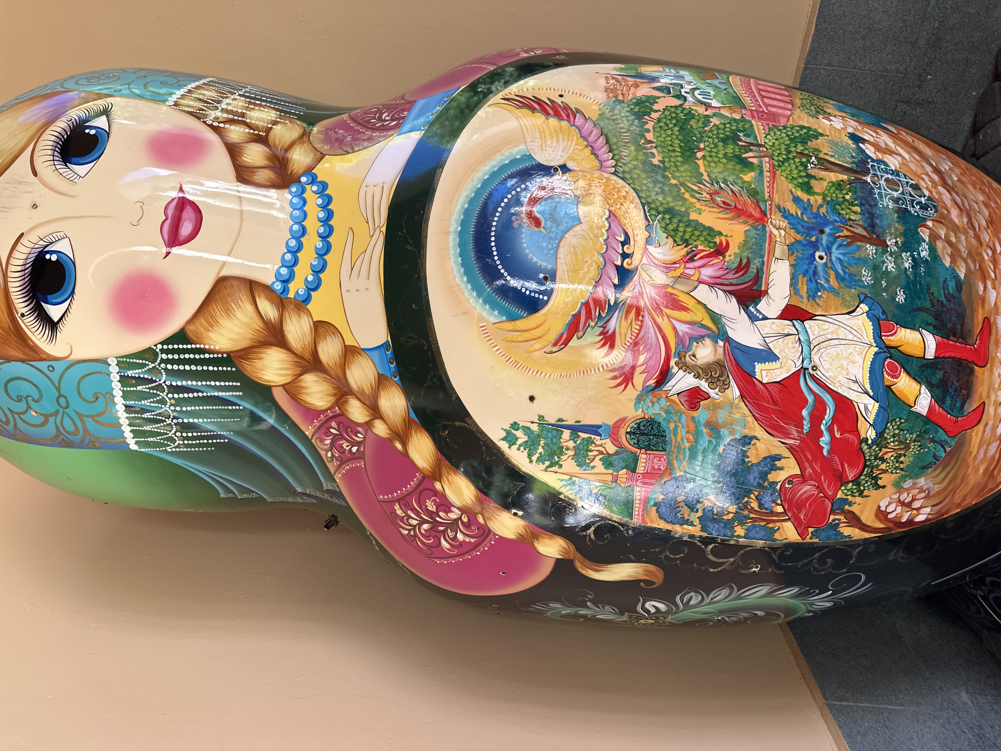 This is a medium shot, eye-level, vertical photograph of a large, intricately painted Matryoshka doll. The doll is positioned against a light peach-colored wall, with a darker tiled floor visible at the bottom. The doll's surface is a canvas for a vibrant and detailed folk art scene.
The upper portion of the doll depicts a woman's face with striking blue eyes, rosy cheeks, and full lips. Her hair is styled in a thick blonde braid that extends down her chest. She wears a blue beaded necklace and a green headscarf adorned with white beadwork and gold patterns.
The middle section of the doll features a circular mural. At its center is a radiant phoenix with yellow and red plumage, its wings spread wide. Below the phoenix, a man in a red cape and white tunic is depicted with his arm outstretched, holding a feather. He is set against a backdrop of lush green trees, colorful buildings with onion domes, and palm trees, suggesting a fantastical landscape. The overall aesthetic is rich in color and traditional Russian motifs. The doll appears to be a stationary object, possibly on display. The location is identified as Estosadok, Russia.