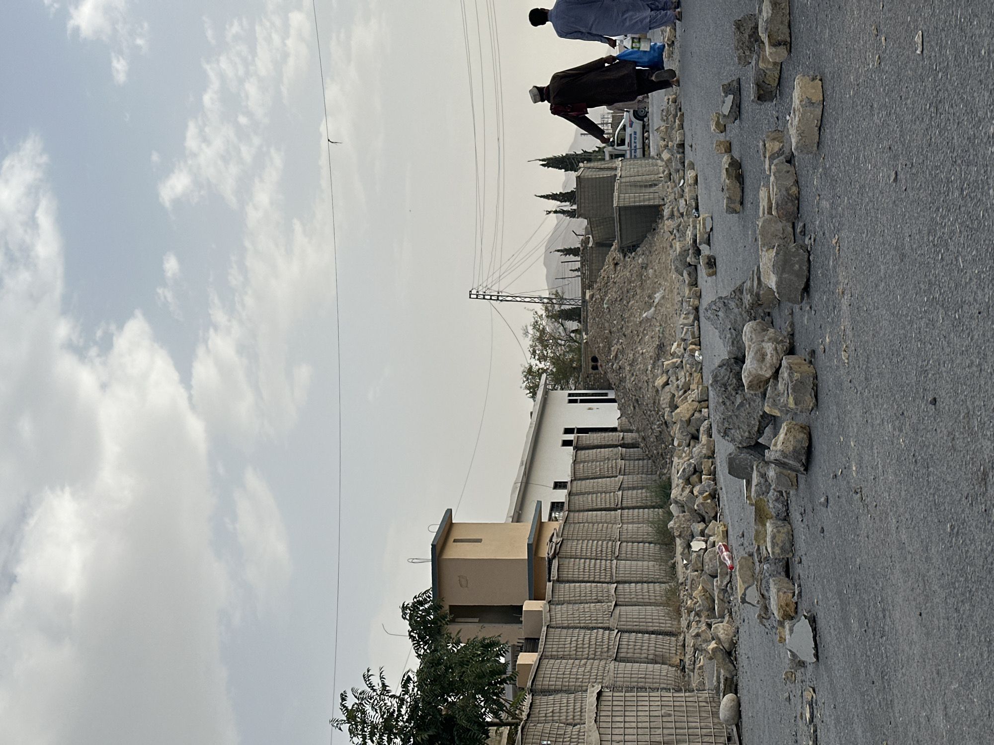 The image depicts a street scene in Quetta City Tehsil, Pakistan, characterized by scattered debris, primarily rocks and building materials, on the asphalt road. A low wall constructed of wire mesh and sandbags lines the left side of the road, with a small concrete guard tower or structure situated behind it. A white building with dark windows is visible behind the sandbag wall. In the background, a police vehicle with "POLICE" written on its side is present. Two men are walking away from the camera on the right side of the road; one is carrying a blue bag and the other a white container. The environment appears somewhat arid with a hazy sky and distant mountains.