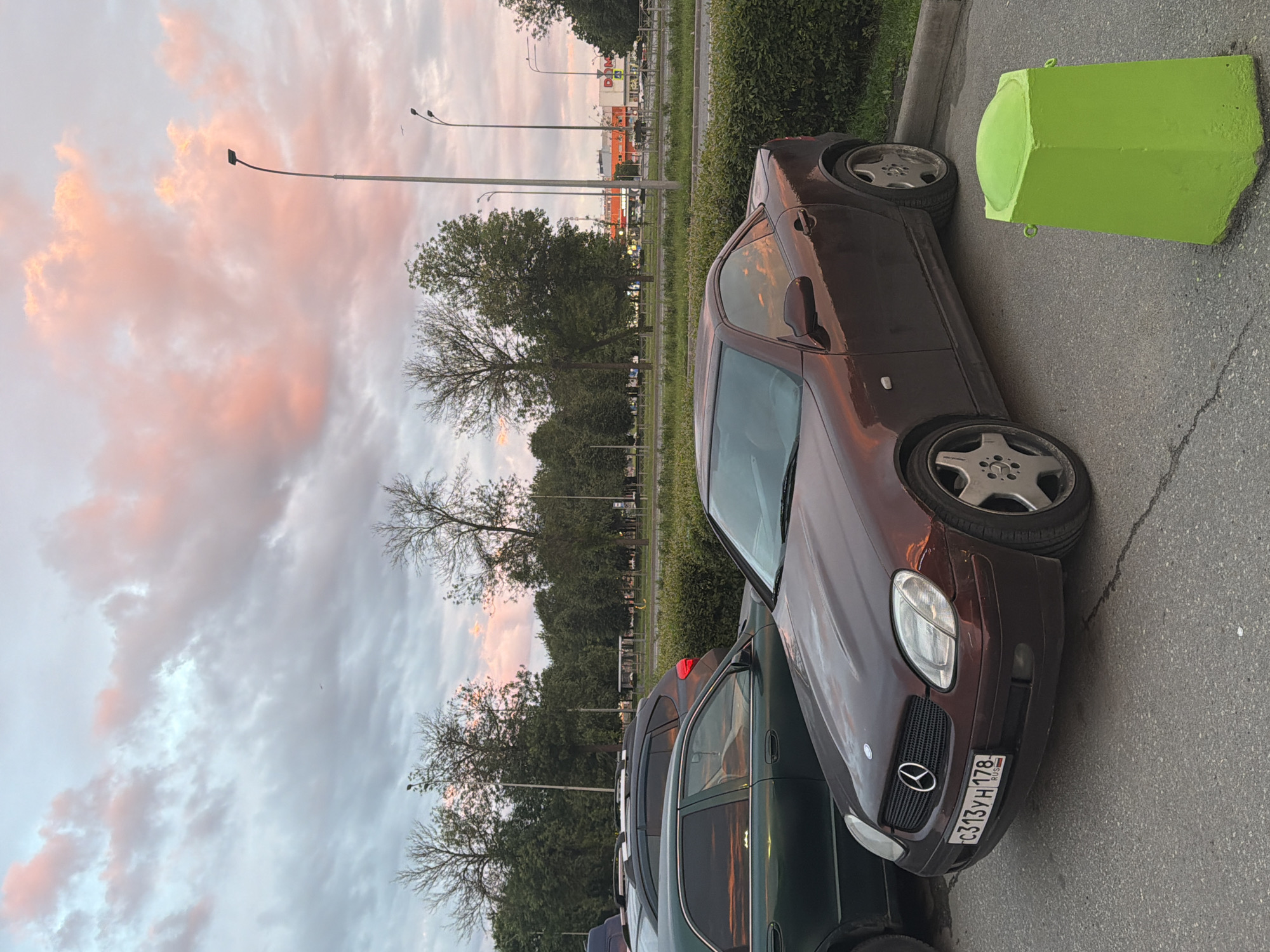 This media file captures an outdoor scene with several cars parked on an asphalt surface. A prominent subject is a dark brown convertible, possibly a Mercedes-Benz, with its front facing the viewer. The car's license plate is visible, displaying the text "C313YH 178 RUS". Next to the convertible, another car, dark green in color, is parked. In the background, there's a row of trees and some buildings, one of which appears to be a supermarket with orange and yellow signage. A bright green concrete barrier is situated near the convertible. The sky is filled with dramatic clouds, tinged with pink and orange hues, suggesting either sunrise or sunset. The overall lighting and the colors in the sky indicate it is either early morning or late evening. The scene appears to be a parking lot or roadside area. There are no people visible in the frame.