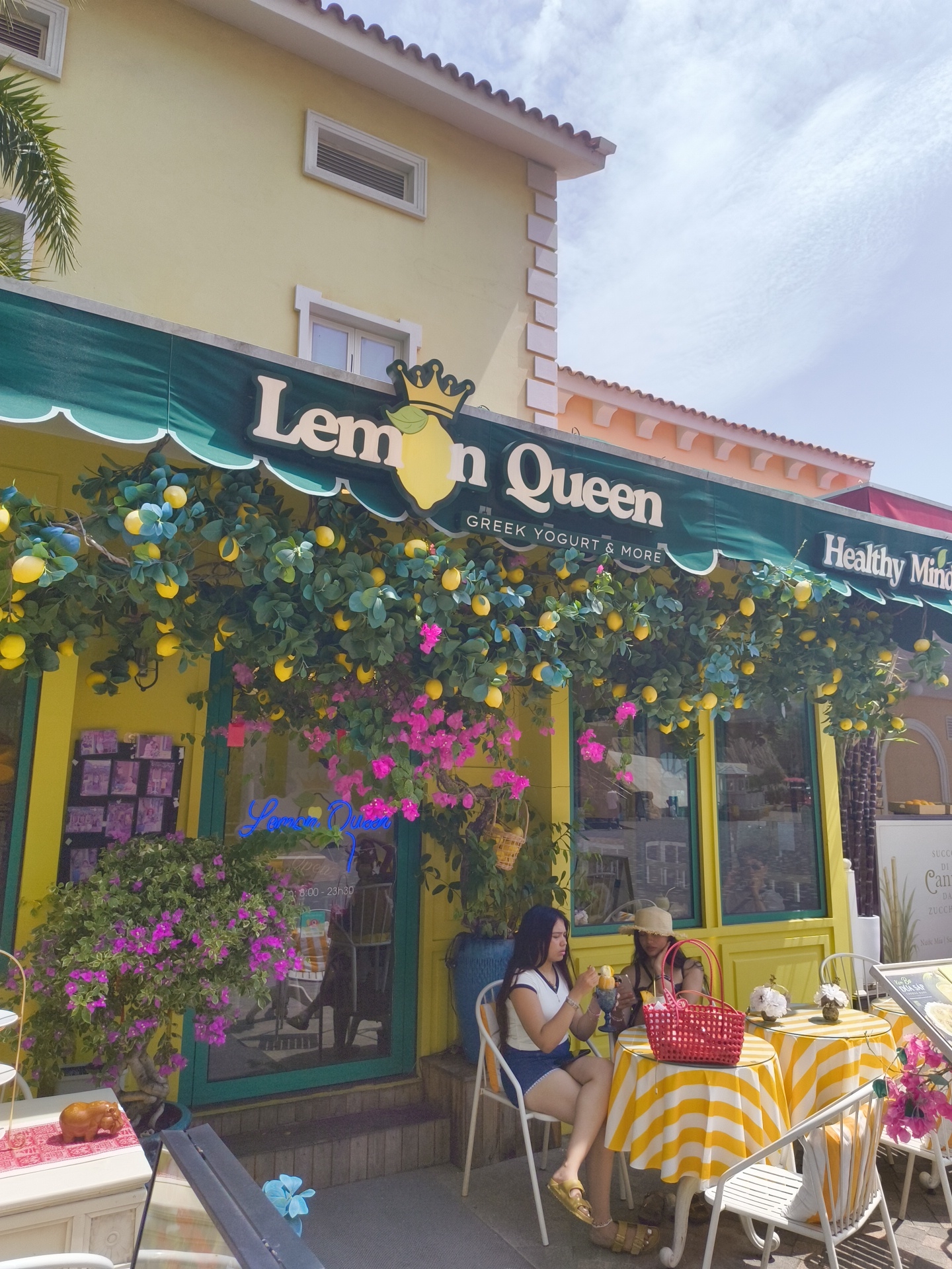 The image shows the exterior of a brightly colored "Lemon Queen" shop in Phú Quốc, Vietnam. The shop's facade is painted yellow with a green awning. Lemon decorations, including faux lemons and pink flowers, are displayed above the entrance and windows. Two women are seated at a small table outside the shop, enjoying what appears to be ice cream or frozen yogurt. The woman on the left is wearing a white top and a blue skirt, while the woman on the right is wearing a straw hat. A red basket sits on their table, which is covered with a yellow and white striped tablecloth. In the background, there is a blue planter with purple flowers and signage for "Lemon Queen" and "Healthy Minds". The overall scene suggests a pleasant outdoor dining or shopping experience.