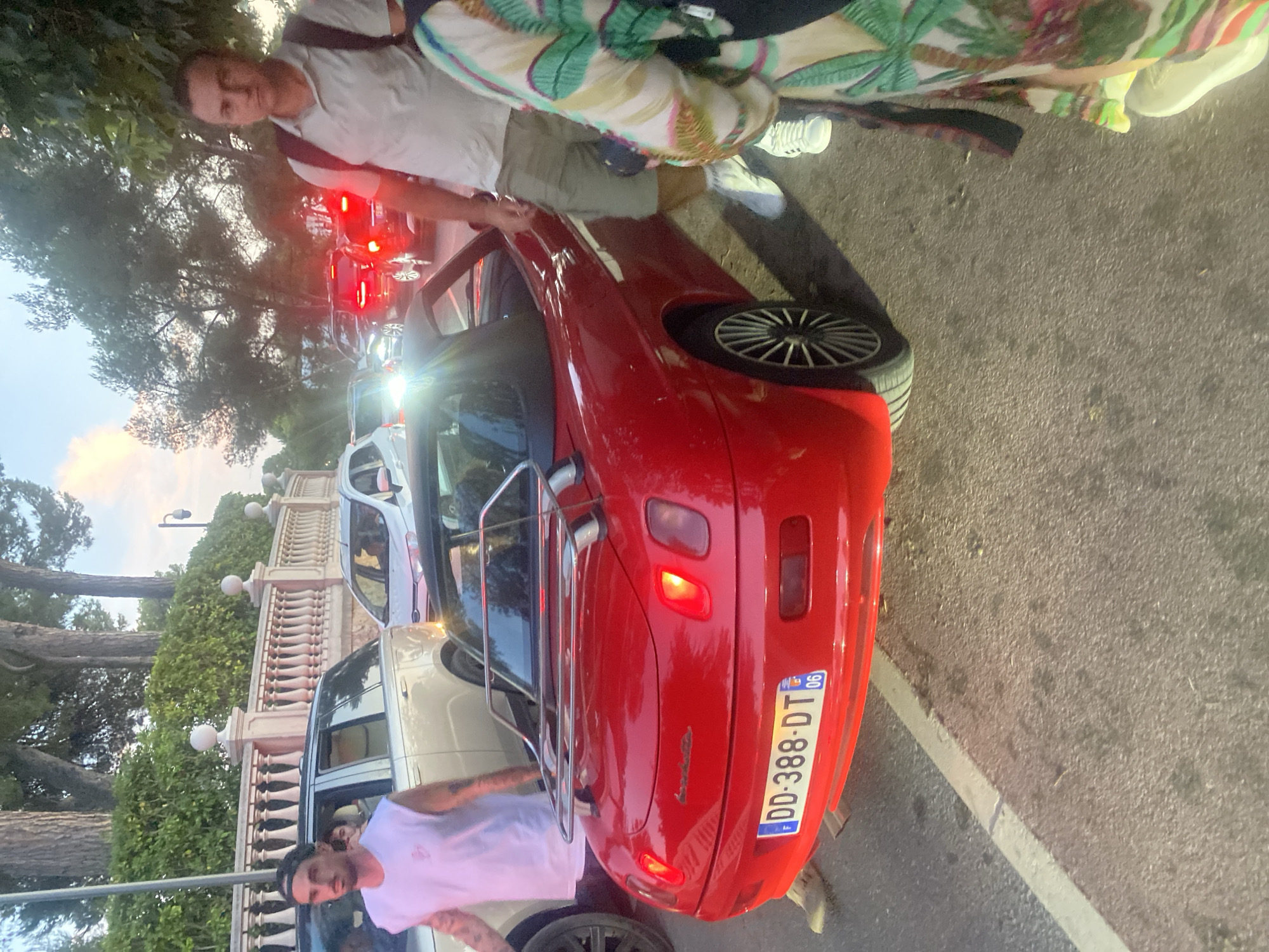 The image depicts a street scene in Antibes, France, likely during the late afternoon or early evening, given the lighting conditions. In the foreground, a bright red convertible sports car is prominently featured, with its license plate reading "DD-388-DT 06". The car appears to be stationary, possibly in traffic, as evidenced by other vehicles visible in the background with their brake lights illuminated.
Several individuals are present. To the left, a man wearing a white sleeveless t-shirt and a baseball cap is walking away from the camera, with a tattoo visible on his arm. He is near a silver SUV. To the right, a man with short dark hair, wearing a light-colored polo shirt and khaki shorts, is looking towards the camera. He has a backpack slung over one shoulder. Another person, whose clothing is partially visible, is standing next to him and wearing a colorful, patterned garment. The background reveals a tree-lined street with a decorative stone wall and balustrade, characteristic of upscale areas. Traffic appears to be congested, with multiple cars visible further down the road. The overall atmosphere suggests a warm climate, typical of the French Riviera.