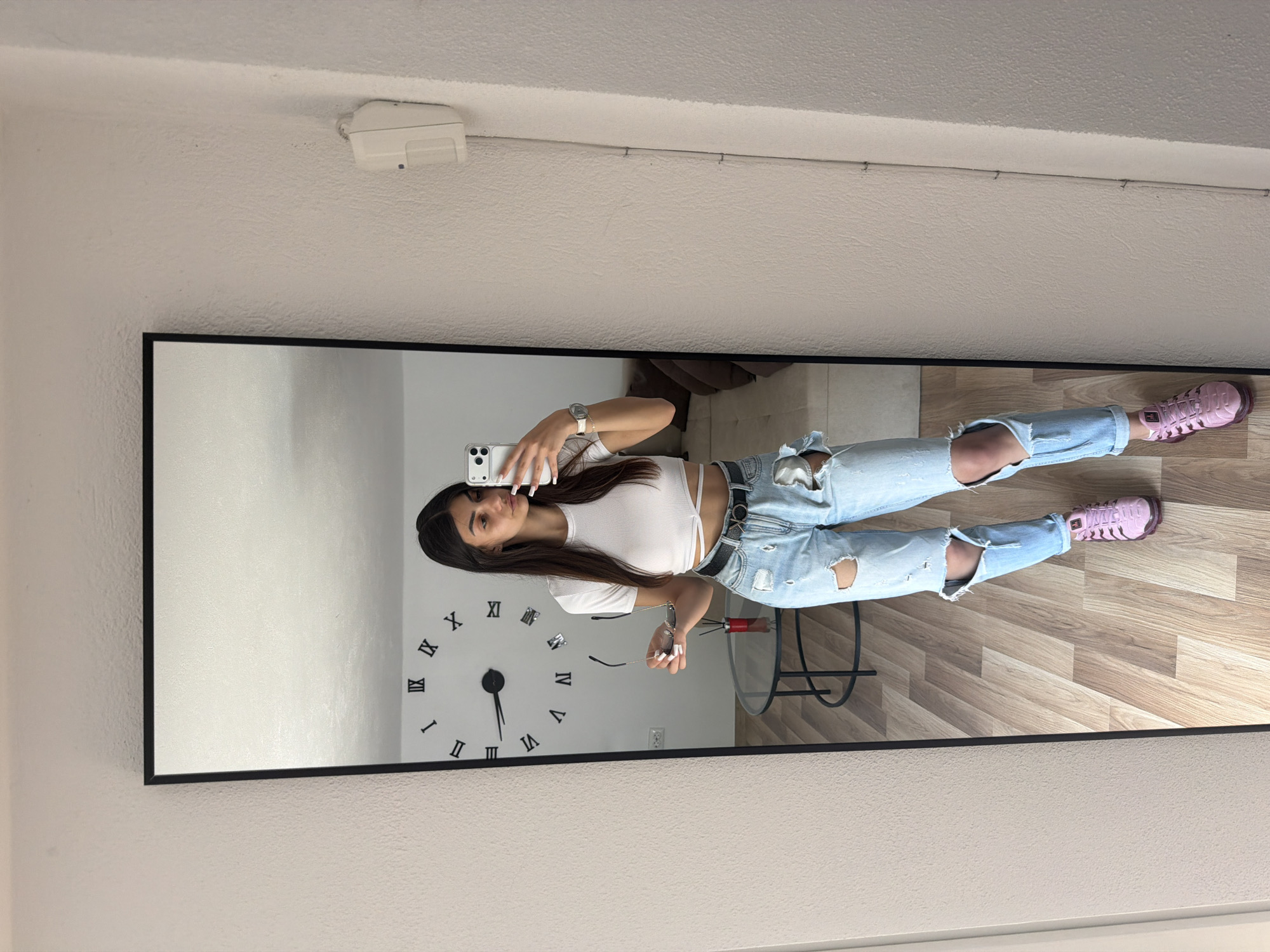 A mirror captures a reflection of a woman taking a selfie. She is wearing a white crop top, ripped jeans, and pink sneakers. A large clock is visible on the wall behind her in the reflection, along with a small table and a section of a couch. The floor is light wood. The mirror itself is framed in black and is hanging on a light beige wall. A white electrical box is visible on the wall above the mirror. The location in the city of Gabrovo, Bulgaria is unknown from the image.