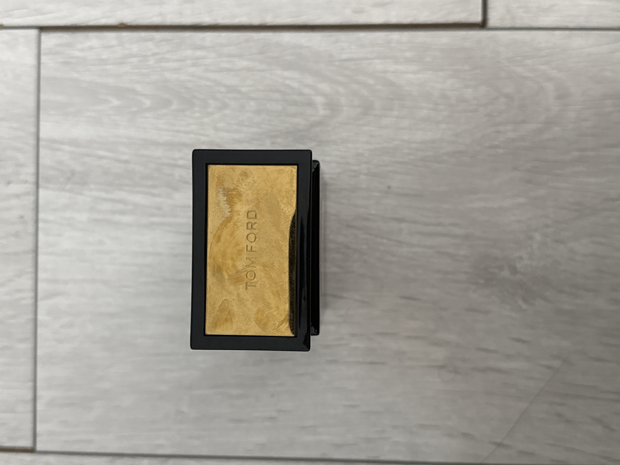 A black rectangular cosmetic compact with a gold lid lies on a light-colored wooden floor. The lid of the compact is embossed with the words "TOM FORD" in capital letters. The background of the image is a wooden floor with a light grain pattern. The image is taken from a top-down perspective. The exact location in Burgas, Bulgaria is not determinable from the image.