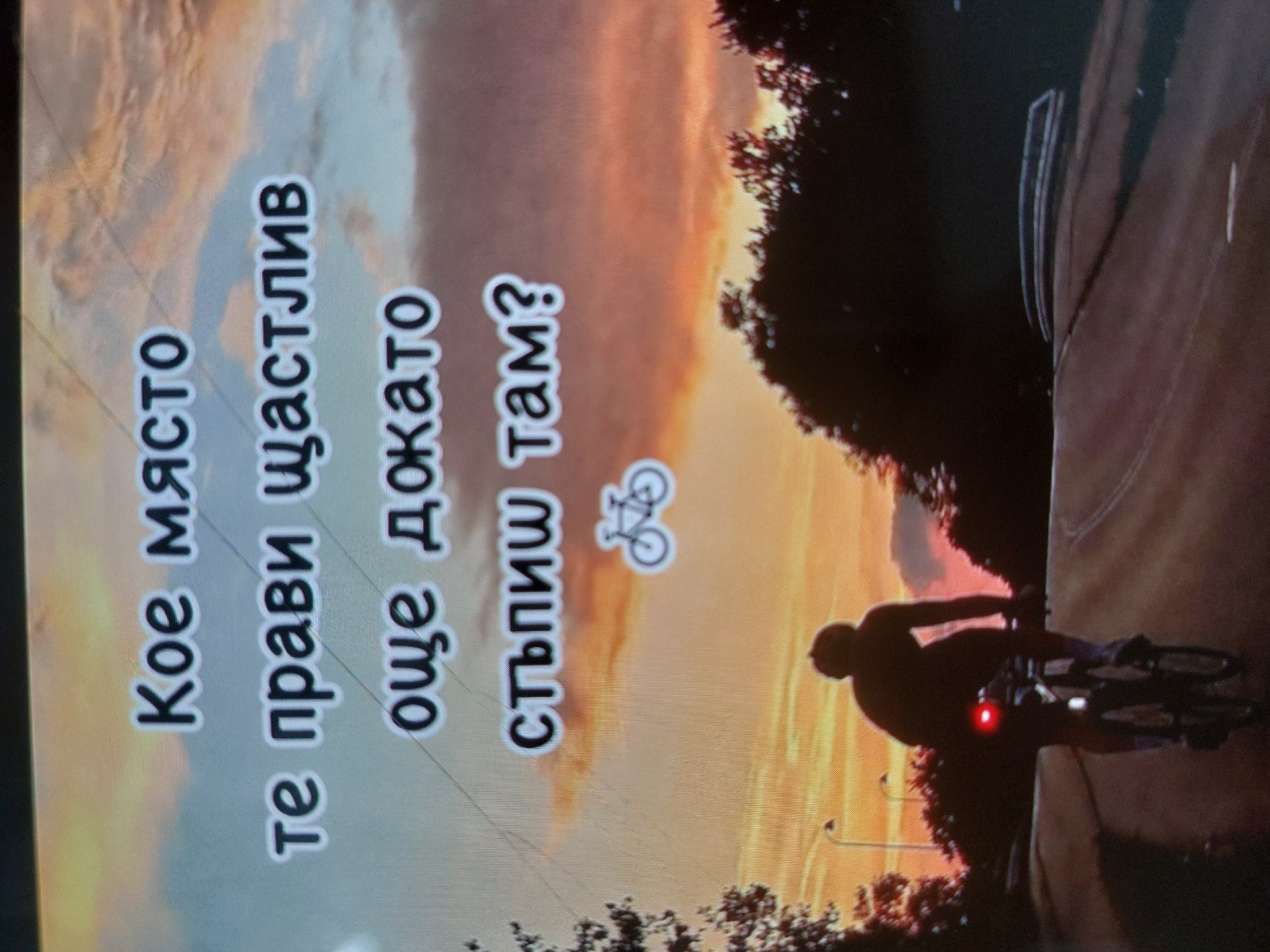 A person is riding a bicycle at dusk. A streetlight is visible in the lower left corner of the image. A road curves to the right. Trees are visible in the foreground. Text in Bulgarian is superimposed on the image. The bicycle has a red rear light. The rider is wearing a helmet.