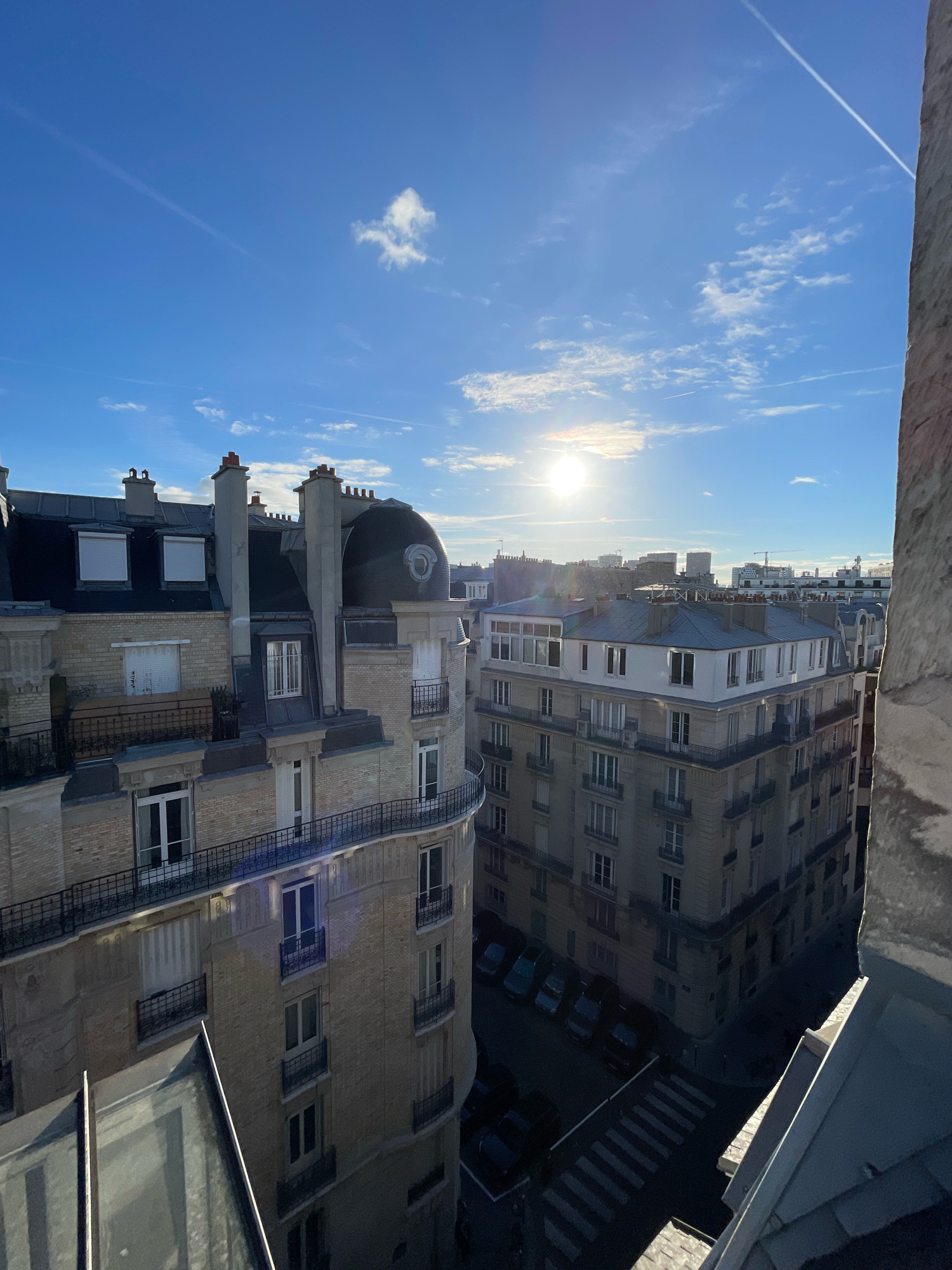 This is an eye-level, medium shot capturing a cityscape in Paris, France, bathed in the warm glow of the morning sun. The image is dominated by the architecture of Parisian Haussmannian buildings, characterized by their stone facades, mansard roofs, and large windows. The sun, low in the sky and partially obscured by wispy clouds, casts long shadows across the scene and creates lens flare.
In the foreground, the roof of a building with a glass-like surface is visible, reflecting the sky and the sunlight. To the left, the ornate facade of another Haussmannian building is prominent, featuring multiple stories with balconies and arched windows. Its light-colored brickwork and dark metal railings add to the classic Parisian aesthetic. To the right, a large, weathered stone structure, likely part of an older building or monument, takes up the edge of the frame, its rough texture contrasting with the smoother buildings.
In the mid-ground, more Haussmannian buildings extend into the distance, their rooftops forming a dense urban landscape. The details of chimneys, dormer windows, and decorative cornices are visible. A curved balcony is noticeable on one of the buildings. Below, a street is visible with parked cars and a pedestrian crossing, suggesting a residential or commercial area.
The sky is a vibrant blue, streaked with thin white contrails from airplanes and scattered, delicate clouds. The overall impression is one of a serene urban morning in Paris, highlighting the city's distinctive architectural style and the gentle morning light. There are no people visible in the image.