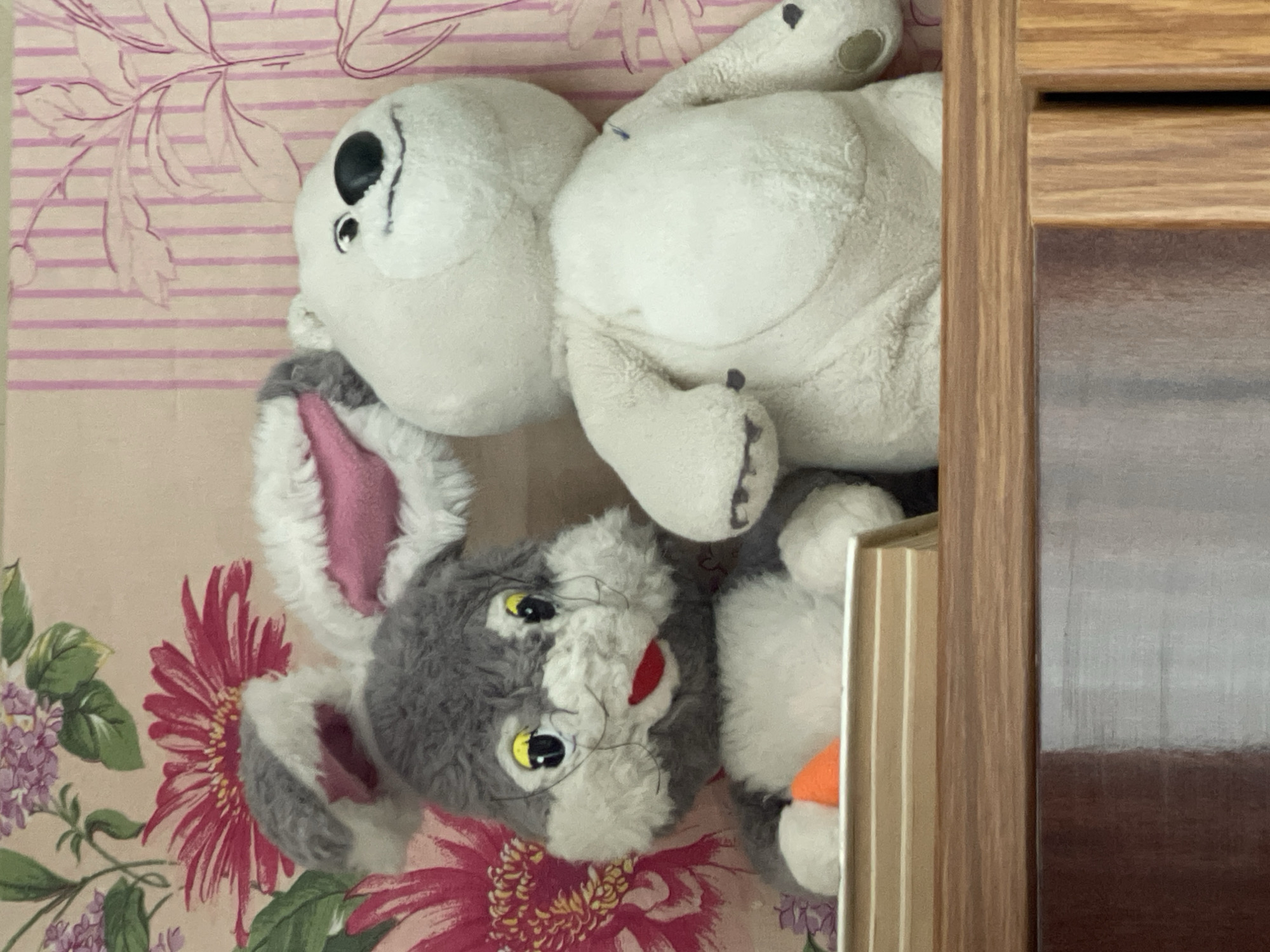 The image shows two stuffed animals: a white bear with black spots and a gray rabbit with pink ears. The bear is lying on its back and the rabbit is positioned below the bear. The background is a floral patterned fabric. The items are near a wooden object. There is no information provided in the image to suggest a location in the city of Zheleznogorsk, Russia.