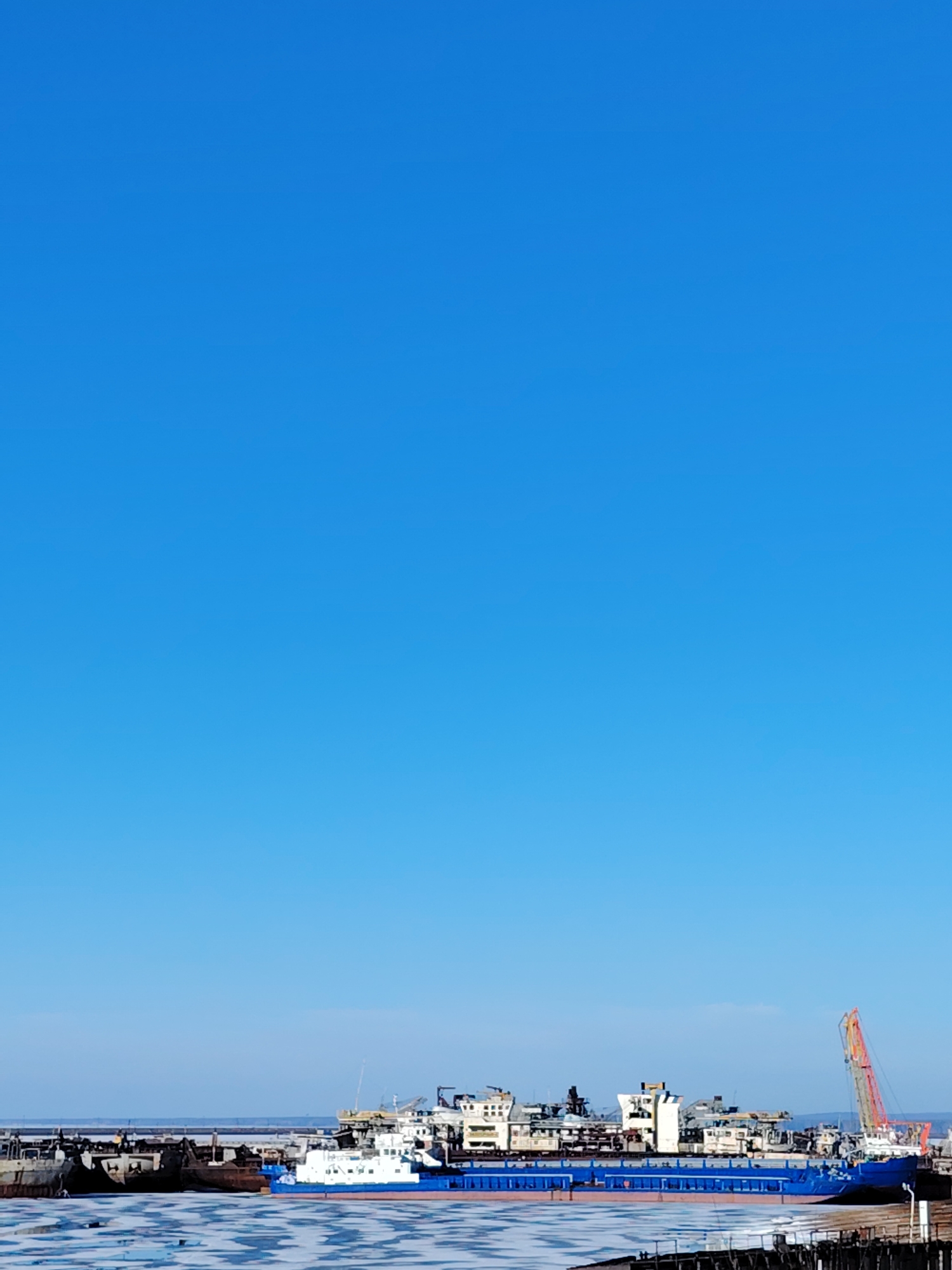 The image depicts a waterfront scene in Chaikovsky, Russia. A large blue and white ship dominates the foreground, docked at a pier or harbor with partial ice cover on the water surface. Various other ships and structures are visible in the background, along with a tall, orange crane. The sky is clear and blue, occupying the majority of the frame.