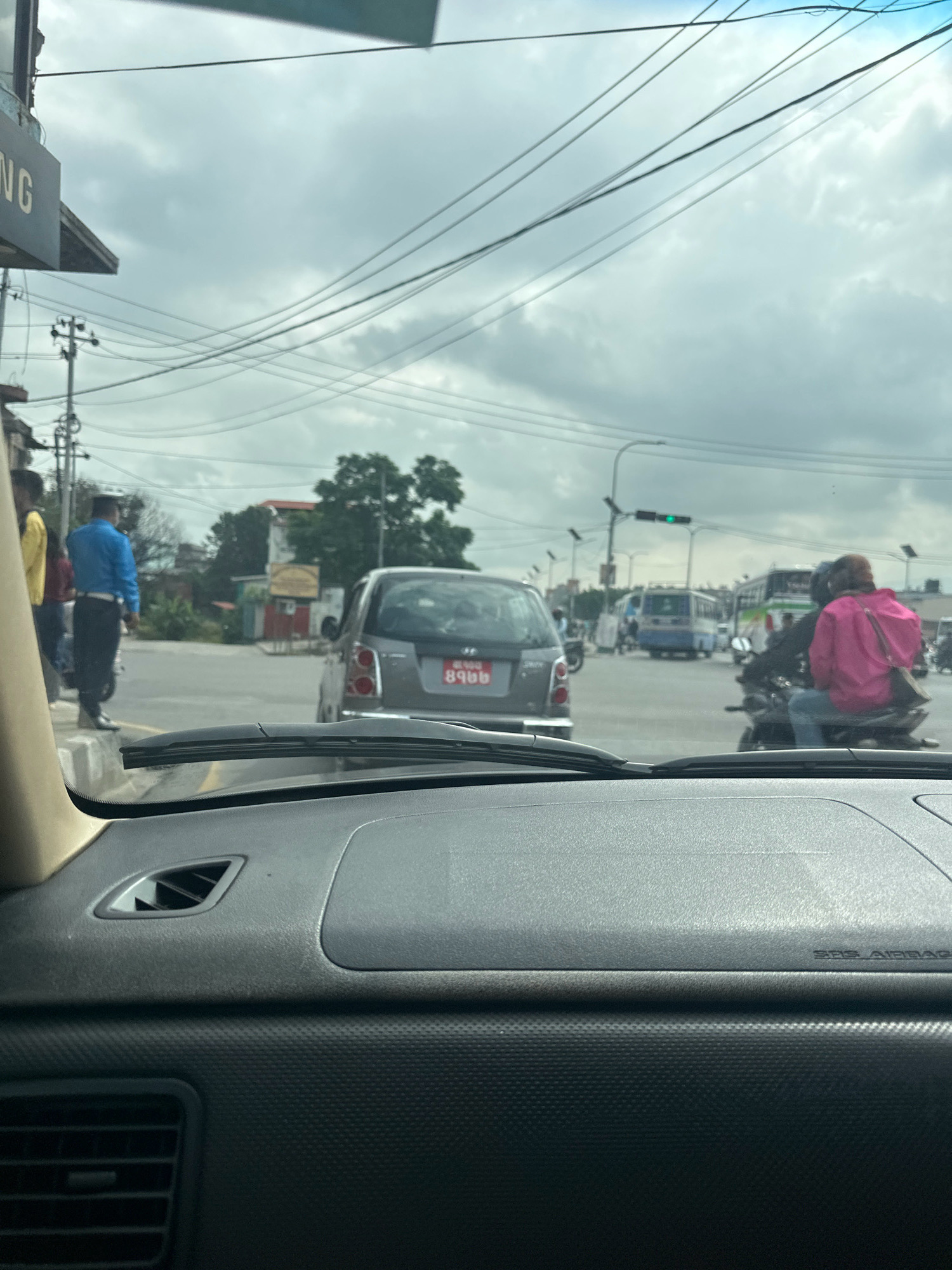 The image shows a view from inside a vehicle, looking out at a busy street intersection in Kathmandu, Nepal. The sky is overcast with clouds. In the foreground, the dashboard of the car is visible. On the street, there is a gray car with a red license plate, a motorcycle with two people on it wearing bright pink and dark clothing, and a person in a blue uniform standing on the sidewalk. Further down the road, several buses, including a blue and white bus, are visible. Traffic lights are present, with one showing green. Electrical wires are strung across the sky, typical of urban areas in Nepal. A sign with "NG" visible is also present on the left side of the image. The scene depicts a typical urban traffic scenario in Kathmandu.