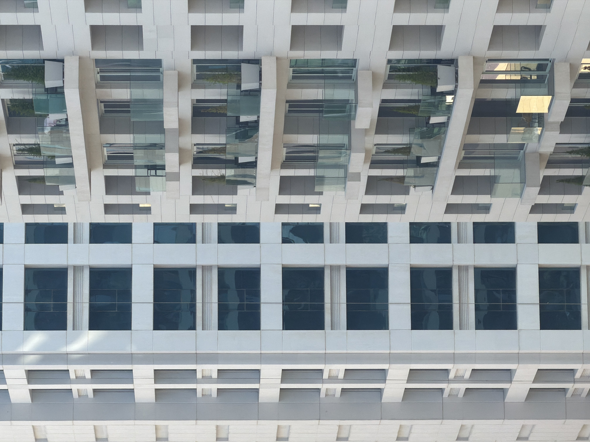 The image shows a close-up of two modern apartment buildings in an urban setting. The building on the left is characterized by its uniform facade of light-colored panels with rows of rectangular windows. The building on the right features a more complex design with balconies on multiple levels. These balconies are equipped with glass railings and some contain potted plants and outdoor furniture. The windows on this building appear to be sliding glass doors. The lighting suggests it is daytime, possibly late afternoon, with sunlight reflecting off some of the glass surfaces. The overall impression is one of contemporary residential architecture.