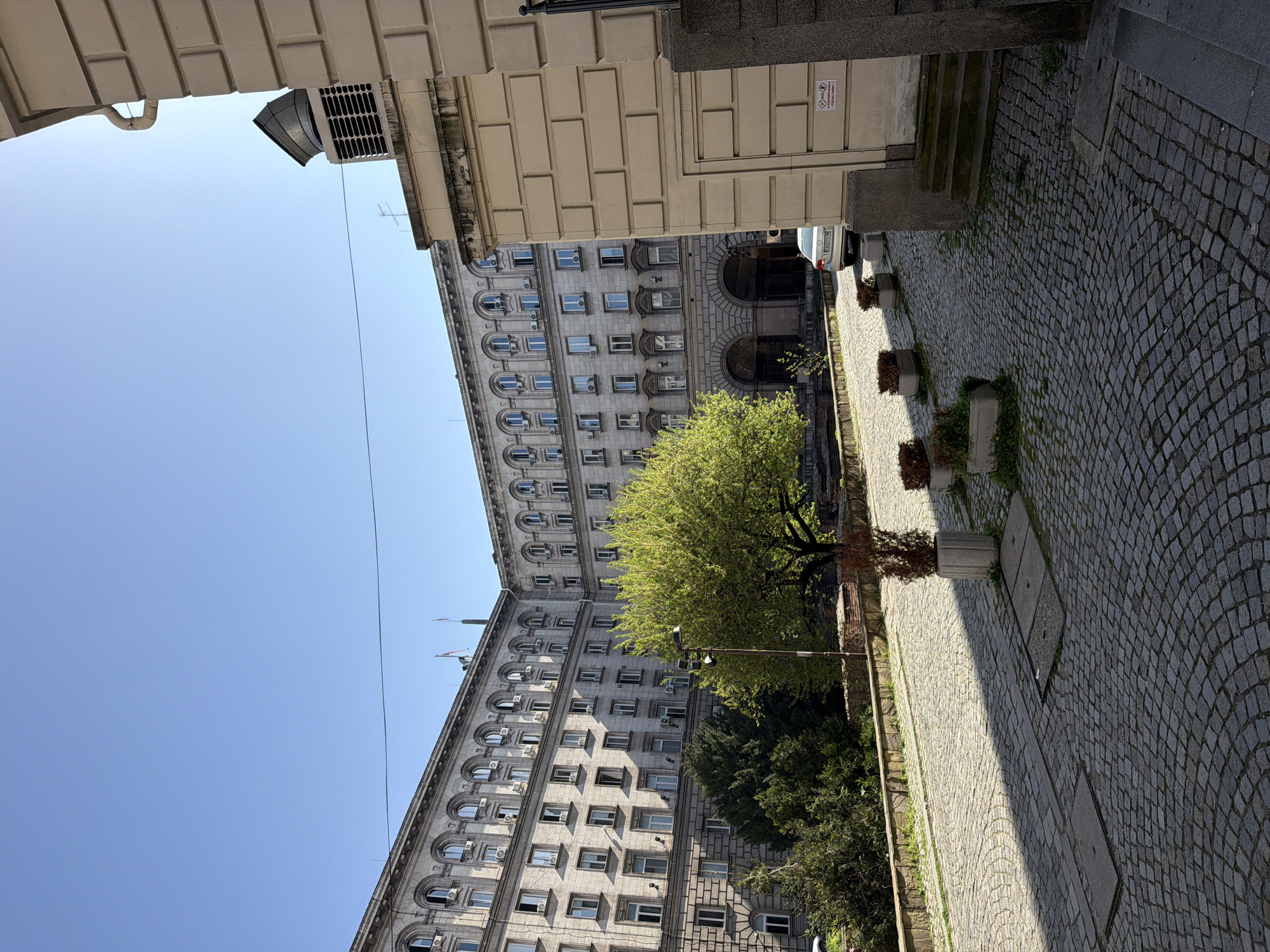 The image captures a sunlit street scene in a European city, specifically Sofia, Bulgaria, characterized by grand architecture and traditional cobblestone paving. No people are visible.
A prominent feature is a large, multi-story building on the left, constructed from light-colored stone or stucco. Its facade is adorned with numerous windows, some arched and others rectangular, many of which have visible air conditioning units. Several flags are just discernible at the top of this building. A lush, vibrant green tree with full foliage stands in the middle ground, casting shadows onto the street below.
The street itself is paved with light-colored cobblestones arranged in an intricate pattern. Along one side, several rectangular concrete planters contain sparse or dry vegetation. Utility access panels or manhole covers are embedded in the pavement. Further down the street, a light-colored car is partially visible.
On the right side of the foreground, parts of another building or structure are seen, featuring prominent brickwork details and forming what appears to be an overhead archway. A white sign with red text is affixed to this structure, bearing the Bulgarian warning: "ЗАБРАНЕНО ПУШЕНЕТО! ПРОДАЖБА И КОНСУМАЦИЯ НА АЛКОХОЛ И НАРКОТИЦИ!" (NO SMOKING! SALE AND CONSUMPTION OF ALCOHOL AND DRUGS!), accompanied by an age restriction symbol.
The clear, bright blue sky above indicates sunny weather, and the distinct shadows cast by the buildings and tree suggest it is daytime, likely mid-morning or early afternoon. The scene conveys a sense of historical urban charm and quietude.