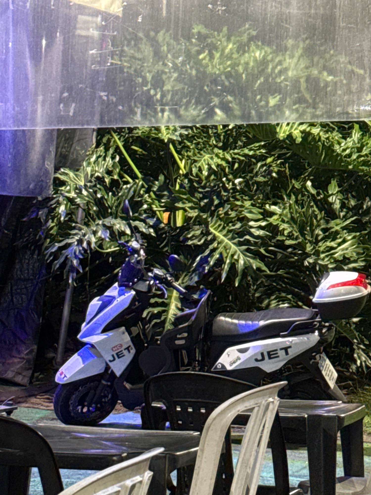 The image depicts a white and blue scooter parked in front of lush green foliage. The scooter has "JET" visible on its side. A white and red top case is attached to the rear of the scooter. The scene appears to be outdoors, possibly in a covered area, as evidenced by a translucent material with some debris hanging above the scooter and foliage. Two black plastic chairs and a table are partially visible in the foreground, suggesting an outdoor seating area. The lighting suggests it is nighttime, with some blue light casting on the scooter. The location is not identifiable within the city of Rio de Janeiro, Brazil.