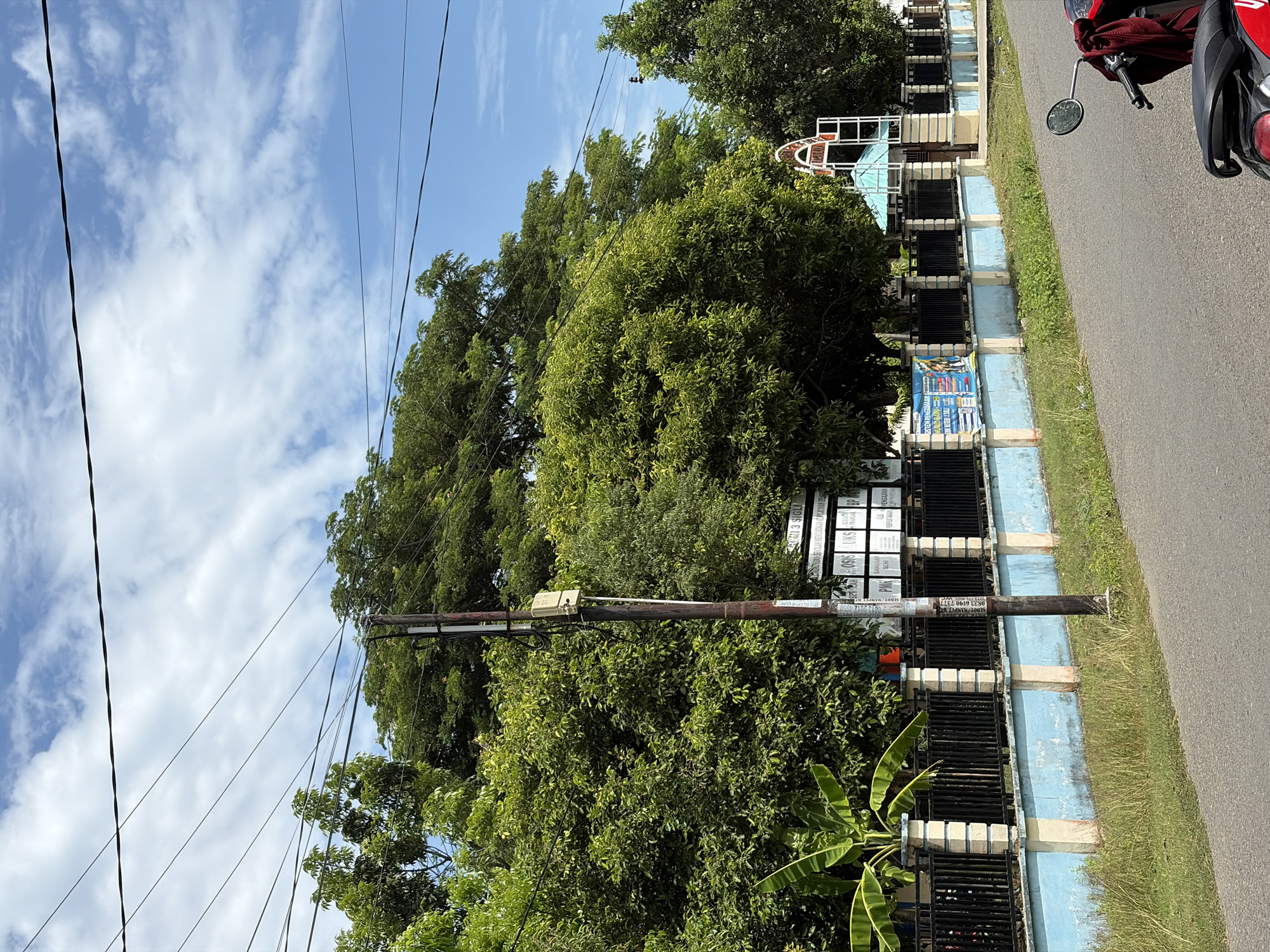The image shows a street scene in Pasi Peukan Baro, Indonesia. A paved road runs diagonally from the lower right corner to the upper left. A motorcycle, partially visible, is parked on the right side of the road. To the left of the road is a grassy verge and a low fence. The fence has a light blue painted concrete base and black metal bars. Behind the fence, there is lush green foliage, including several large trees and what appears to be a banana plant. A wooden utility pole with some electrical equipment attached stands prominently in the foreground, near the fence. Further back, a building with a light blue awning or roof is visible, along with a white arched structure. Several signs are mounted on the fence, one of which appears to have text in Indonesian. Overhead, power lines are strung across a blue sky with scattered white clouds.