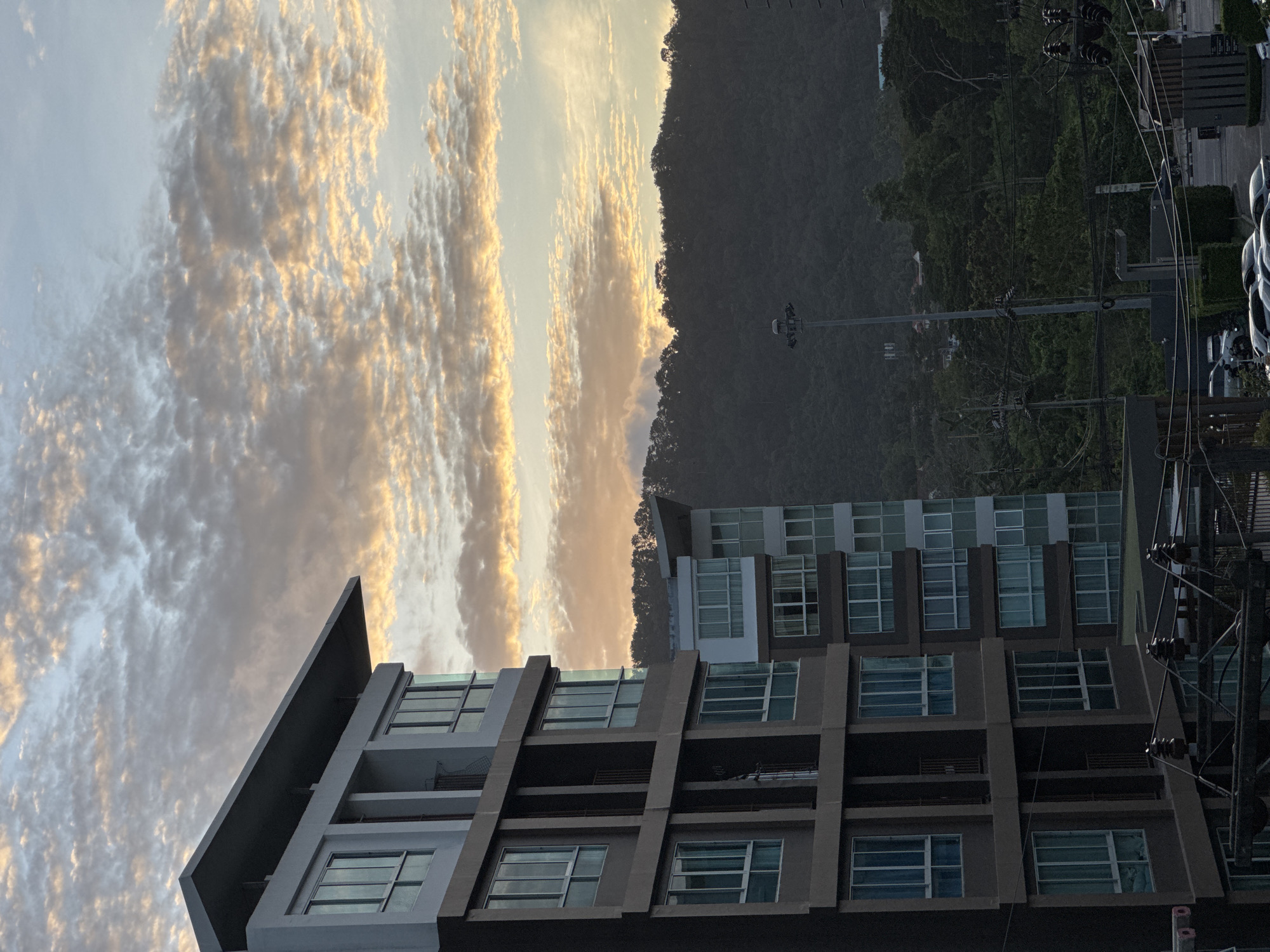 The image, captured in Kathu, Thailand, depicts a modern apartment building against a backdrop of a tree-covered hillside and a sky filled with patterned clouds. The building, characterized by its dark brown and white facade and numerous windows, dominates the left side of the frame. To its right, a second, lighter-colored apartment building is partially visible. The sky displays a dramatic display of cumulus clouds, illuminated by the warm hues of what appears to be either sunrise or sunset, casting a golden glow. In the distance, the dense green foliage of a hill forms a natural boundary, with hints of houses nestled among the trees. The foreground is occupied by a street with parked cars and utility poles with visible electrical wires. The overall scene suggests a contemporary urban environment seamlessly integrated with a natural landscape, bathed in the soft light of dawn or dusk.