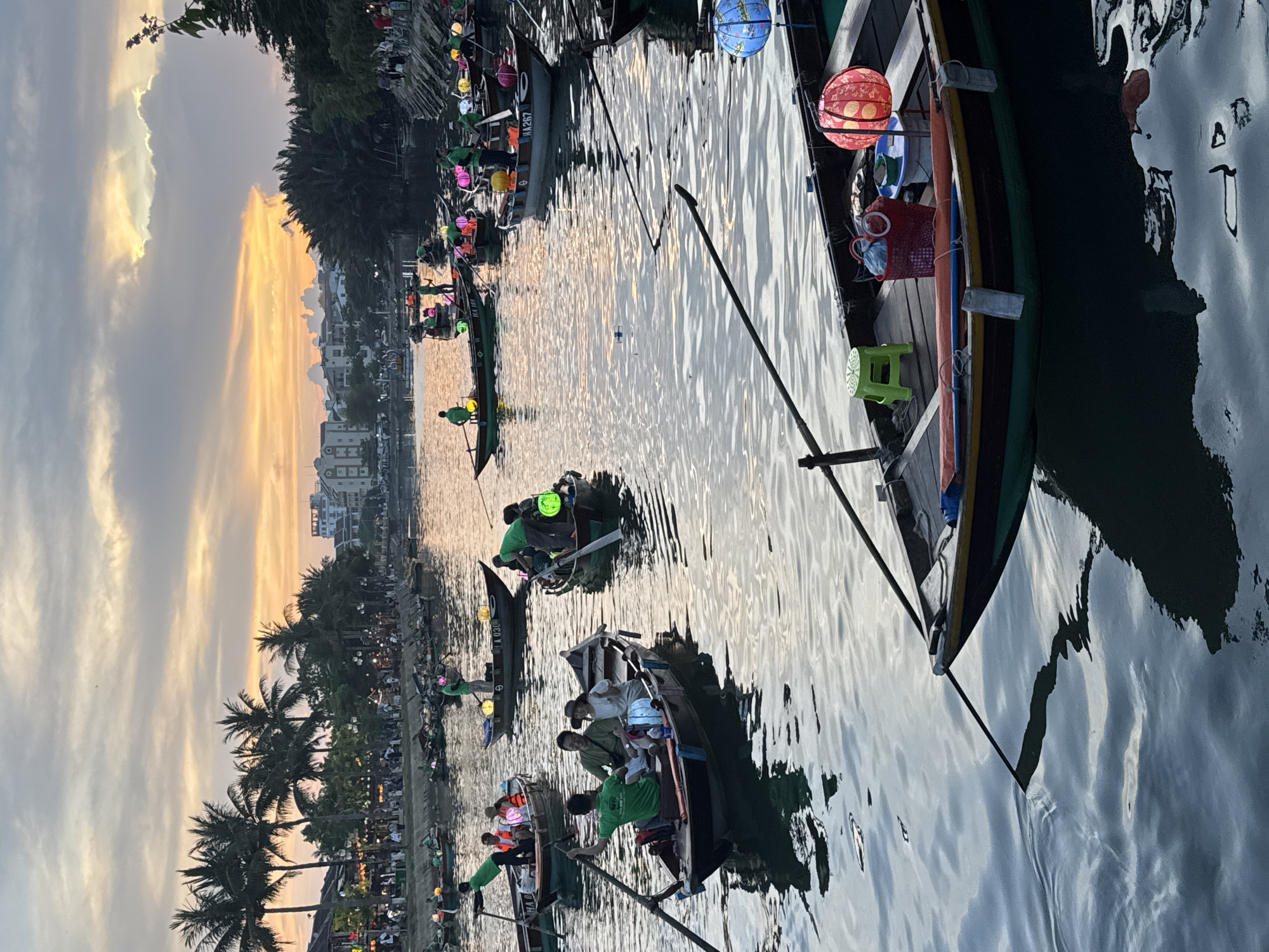 The image shows a waterfront scene in Hội An Ward, Vietnam, at dusk. Several boats with passengers are on the water; some boats have lanterns. People are on the shore under palm trees, and buildings are visible in the background. The sky is partly cloudy with a sunset glow.
