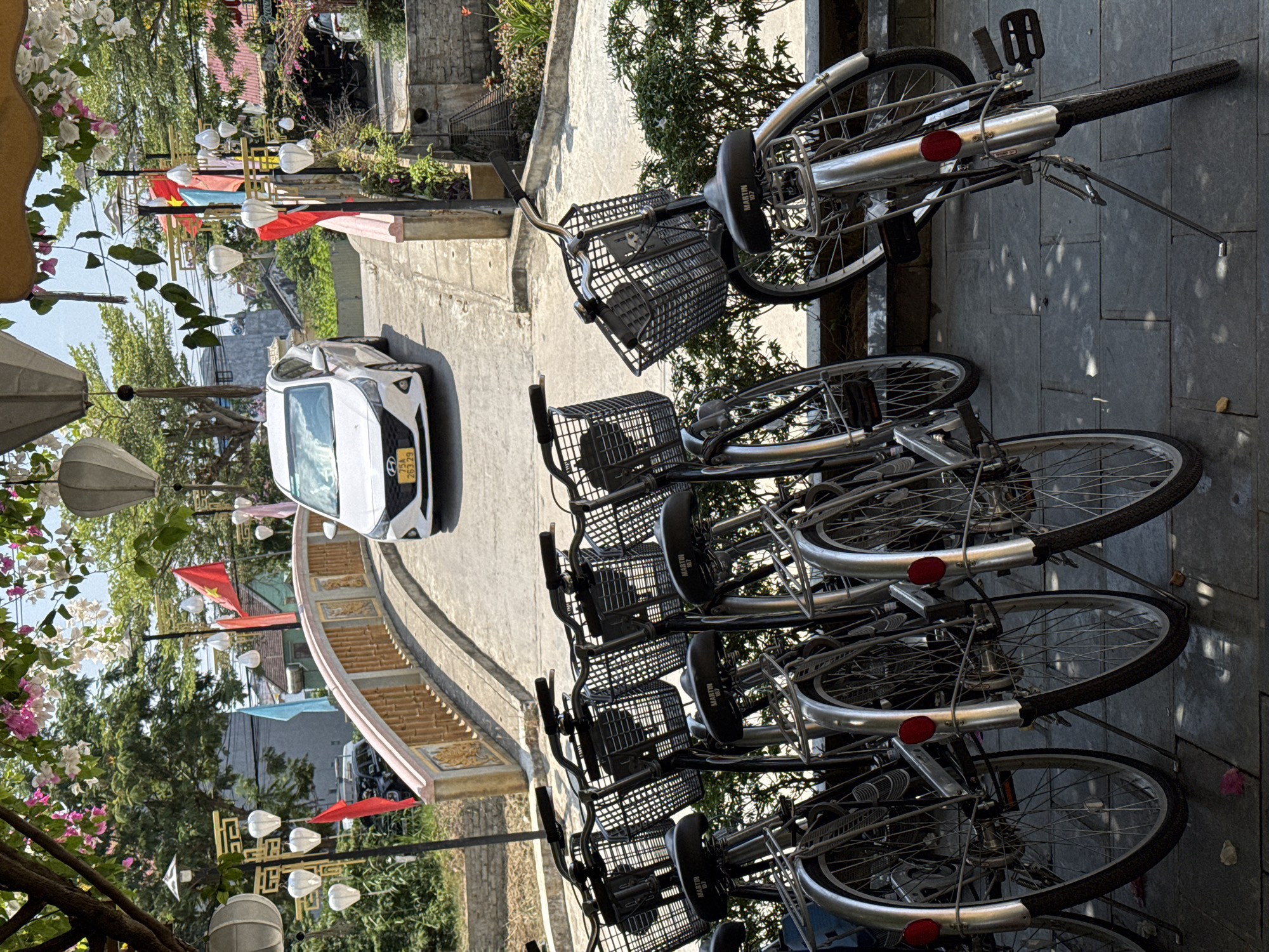 The scene shows a row of bicycles parked on a paved surface in Hội An Ward, Vietnam. The bicycles are black and silver, with baskets on the front. A white car is parked on a nearby street. There is a bridge with yellow ornate trim and red flags on poles. There are trees with flowering vines and light posts with lamps.