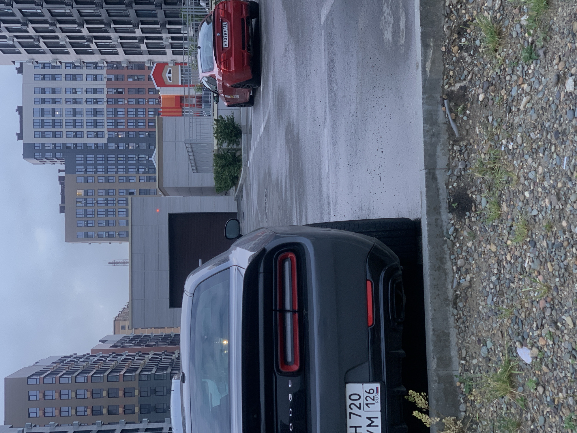 The image shows a parking lot in a modern urban environment, likely in Stavropol, Russia, due to the visible license plate with "RUS". In the foreground, the rear of a dark grey Dodge Challenger is visible, parked facing away from the camera. The car has a distinctive taillight design and a spoiler. To the right and further back, a red BMW M3 coupe is parked, also facing away. The cars are situated on a wet asphalt surface, indicating recent rain. Surrounding the parking lot are several tall, contemporary apartment buildings with varied architectural designs and color schemes, typical of a newly developed urban area. The sky is overcast. The scene is devoid of any people.