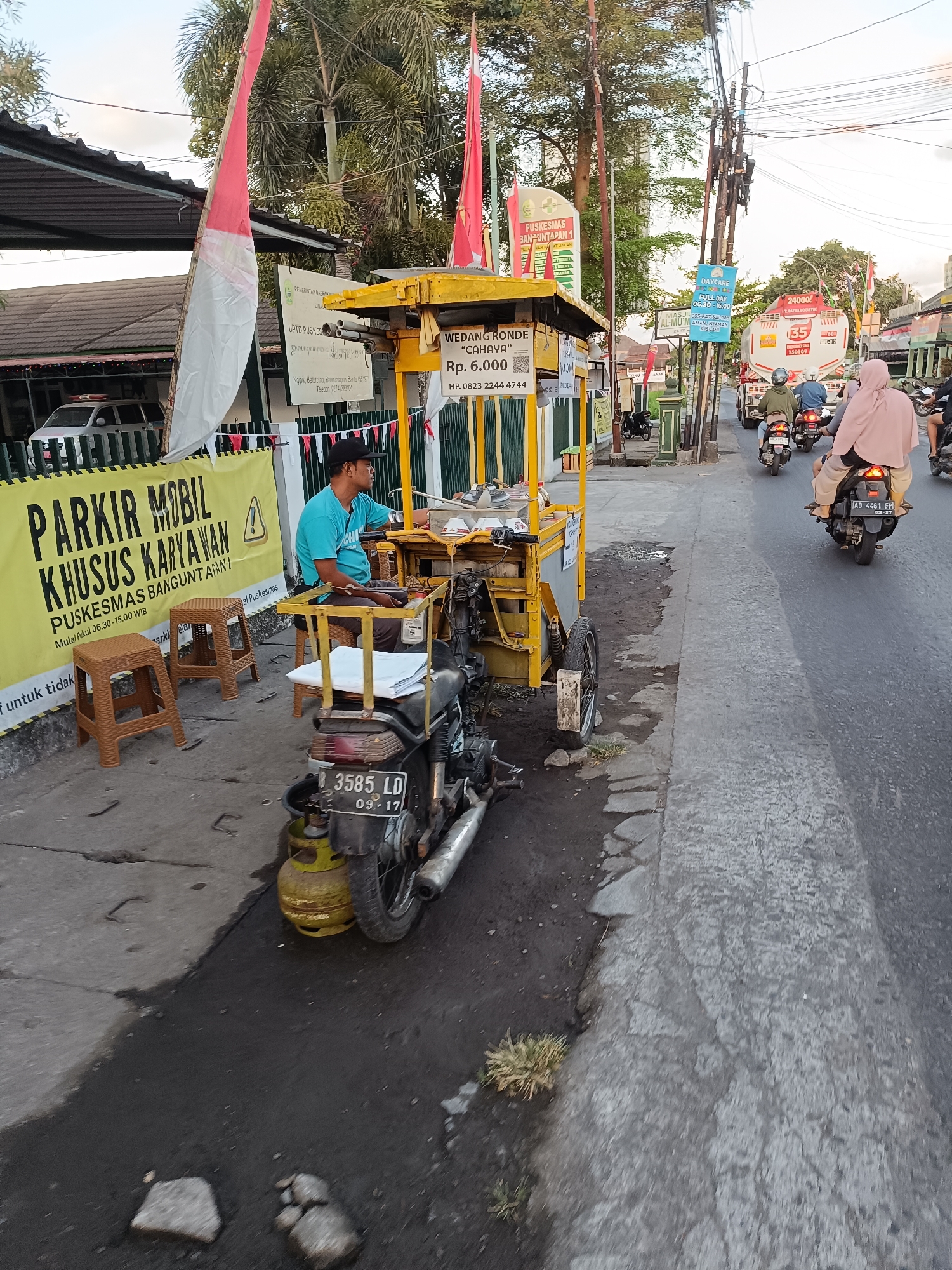 A man wearing a blue t-shirt and a black baseball cap is sitting on a stool next to a yellow food cart that is attached to a motorcycle. The food cart has "WEDANG RONDE" and "Rp 6.000" printed on it. The motorcycle has a gas tank attached to the back. There are several red and white flags on poles behind the food cart. A banner with yellow and black text is hanging on a fence to the left of the food cart. The banner reads "PARKIR MOBIL KHUSUS KARYAWAN PUSKESMAS BANGUNTAPAN". The scene appears to be on a street in Banguntapan, Indonesia, with traffic visible in the background, including several motorcycles and a truck. The pavement is wet in some areas.