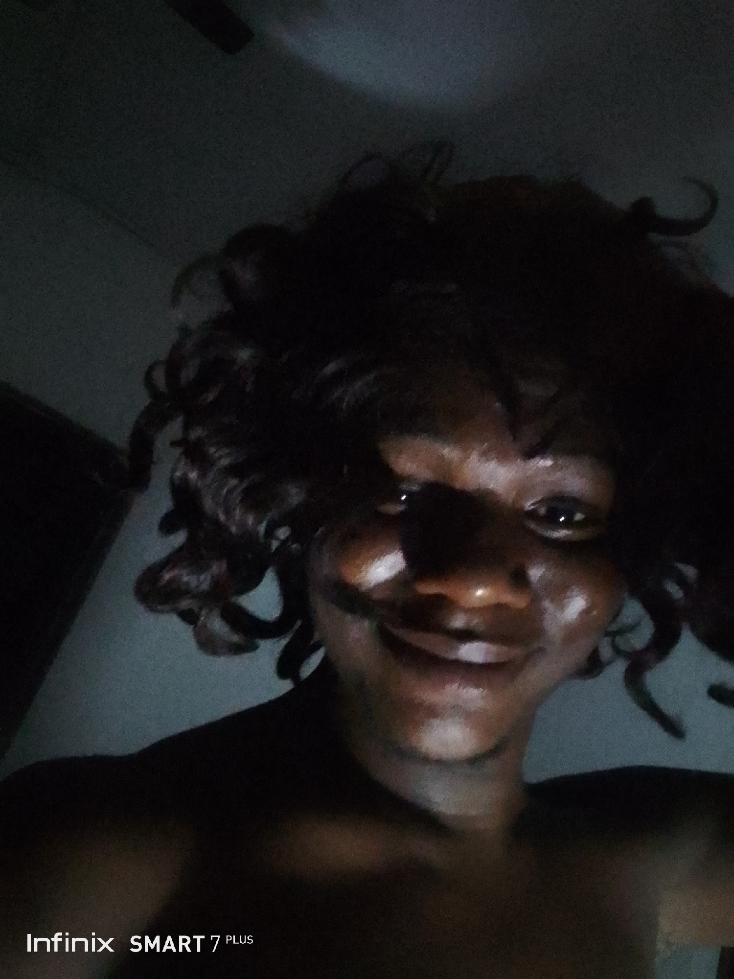 The image displays a low-light, close-up selfie of a person with dark skin and dark, curly hair. The individual is positioned looking upwards towards the camera, with a noticeable smile on their face, suggesting a cheerful or relaxed mood. The lighting is dim, casting much of the scene in shadow, but highlights are visible on the person's forehead, cheeks, nose, and lips, indicating a light source directed towards the face.
The person appears to be shirtless or wearing dark attire that blends into the dark surroundings. The background is largely indistinct due to the low light, but hints of an indoor setting are present; a faint outline resembling a ceiling fan can be seen in the upper left portion of the image. The overall scene suggests the photo was taken at night or in an interior space with minimal illumination.
In the bottom left corner, the text "Infinix SMART 7 PLUS" is visible, identifying the device used to capture the photograph.