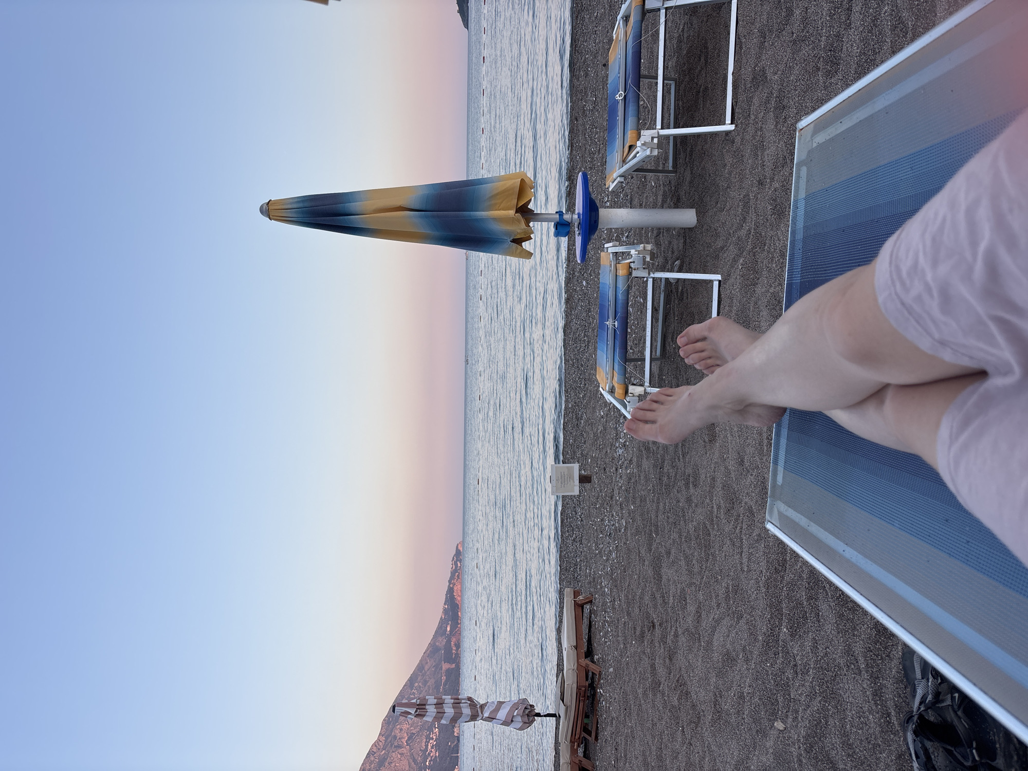 The image depicts a first-person perspective of someone relaxing on a lounge chair on a pebble beach in Bečići, Montenegro. The scene is set at dusk, with the sky displaying a gradient of soft blues and pinks. The calm sea stretches out to the horizon, with a distant coastline and mountains visible on the left.
In the foreground, the legs and feet of the person are visible, resting on a blue and white striped lounge chair. To the left of the person's feet, a pair of black sandals and a dark bag are partially visible.
Several beach chairs and closed umbrellas are arranged on the beach. One large umbrella, with blue and yellow stripes, is centrally located, standing beside a small blue table. Another umbrella, with white and brown stripes, is positioned further to the left, partially obscuring a row of beige lounge chairs. A white sign is also visible on the beach. The pebbles on the beach are dark and small, with some larger stones mixed in. The water appears tranquil and reflects the soft hues of the sky.