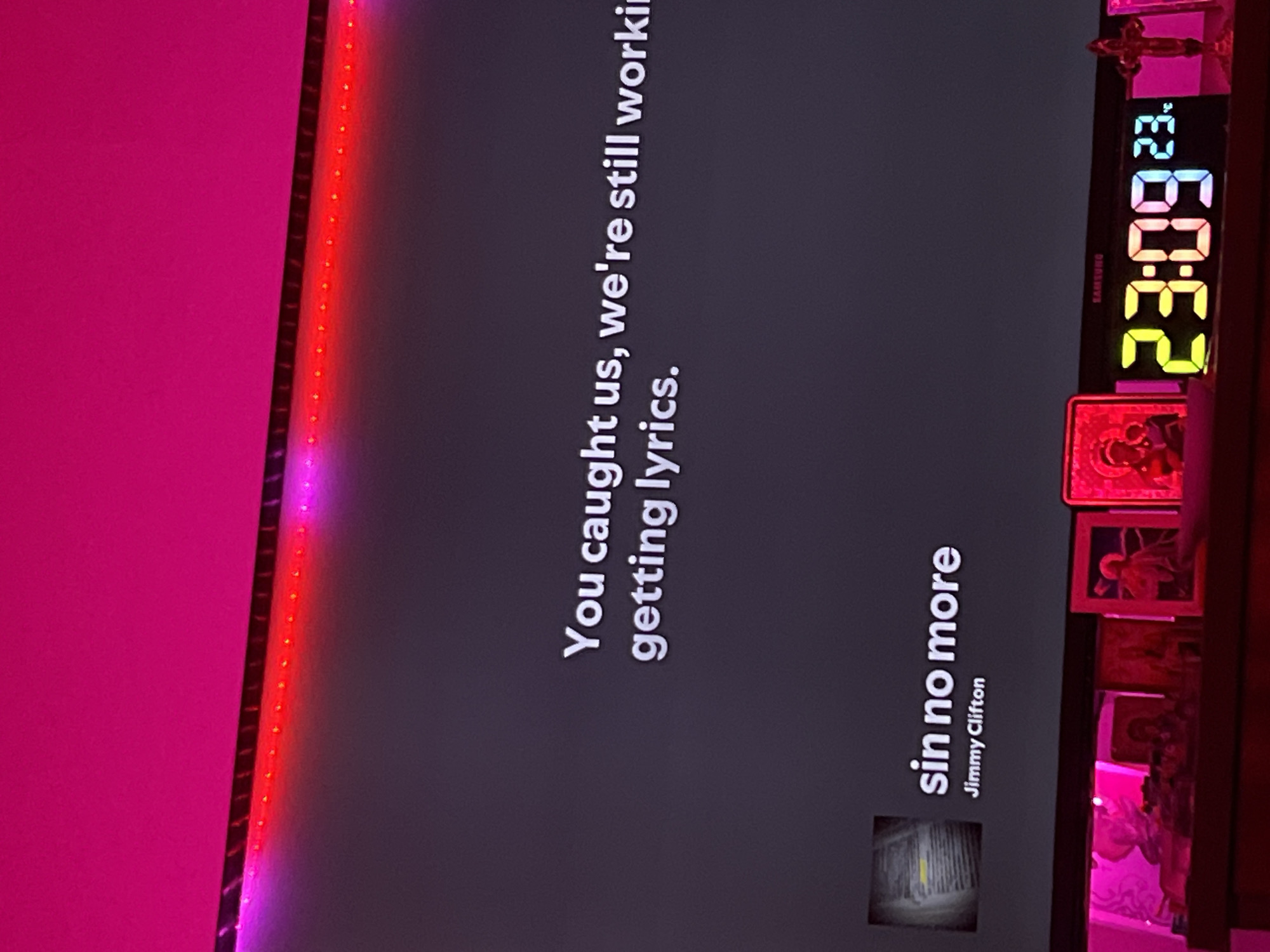 The image displays an indoor night scene with a television screen as the focal point, featuring active LED strip lighting and various decorative items on a surface below.
At the top, a wall is illuminated by a diagonal LED strip, transitioning from a magenta hue on the left to a vibrant red towards the right.
The central and dominant element is a dark grey screen displaying a message in white text: "You caught us, we're still working getting lyrics." Below this message, on the lower left of the screen, album art (appearing as abstract pages with highlighted text) is shown next to the song title "sin no more" and artist "Jimmy Clifton".
Below the screen, a dark, reflective surface (possibly wood) supports several objects. These include multiple framed pictures or religious icons, one prominently glowing with a magenta hue on the far left. A smaller, golden crucifix stands upright on the far right. Between these items, a multi-colored digital clock displays "23:09" in large, segmented numbers (yellow, orange, red, blue), with smaller blue digits showing "23." to its right. The "SAMSUNG" brand name is faintly visible above the clock. The objects on the surface are bathed in a magenta ambient light, reflecting the room's illumination.
There are no individuals present in the image. The visual content provides no information to determine the geographical location, such as the city of Debelt, Bulgaria.