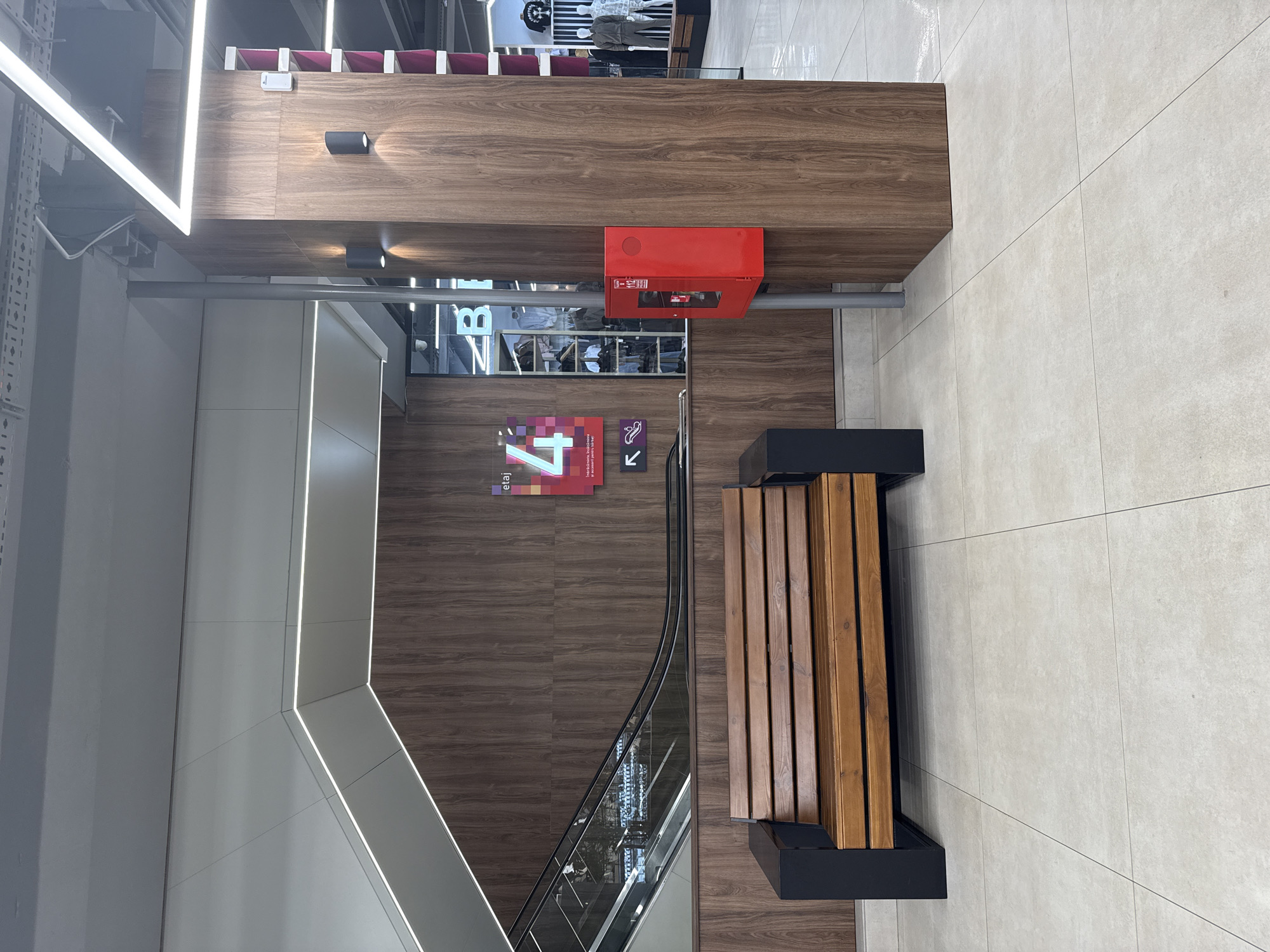 A wide shot captures an interior scene, possibly a shopping mall or public building, featuring modern architecture and finishes. In the foreground, a wooden bench with dark metal accents sits on a tiled floor. Behind the bench, a wall is paneled with dark wood veneer. To the left, an escalator with glass railings ascends along a similarly paneled wall. On the right, a large, dark wood-clad pillar supports overhead lighting. Attached to the pillar is a bright red fire extinguisher cabinet. Further in the background, the scene opens up to reveal a retail environment with mannequins dressed in casual attire and shelves displaying merchandise. The overall impression is one of a clean, contemporary commercial space.