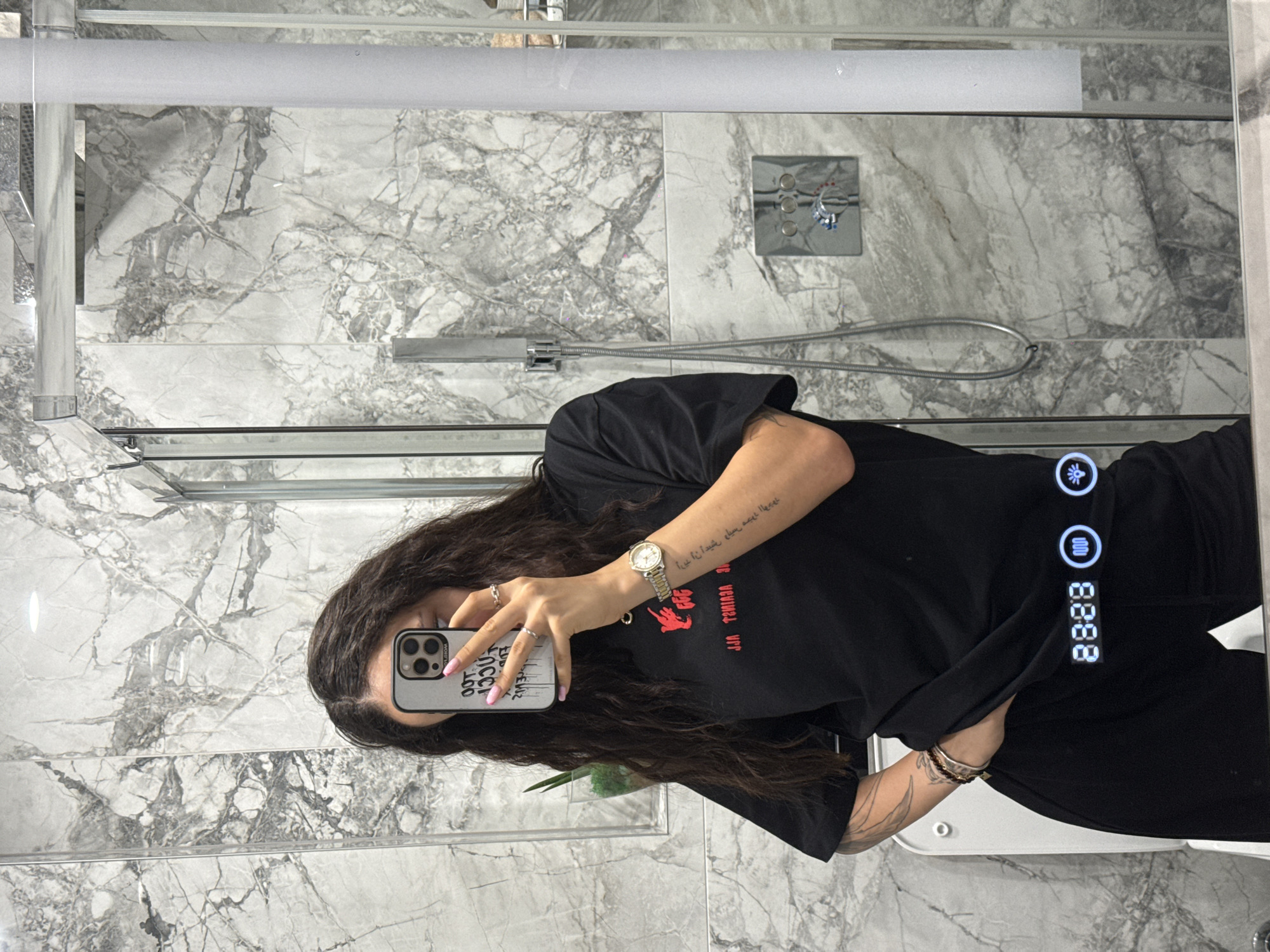 The image shows a woman taking a mirror selfie in a bathroom. She is wearing a black t-shirt and black pants, and holding a phone in her right hand. The bathroom has marble walls and a glass shower enclosure. A digital display on the mirror shows the time "88:88" and two illuminated icons. A shower head and faucet are visible on the wall. There is no discernible location in Shumen, Bulgaria.