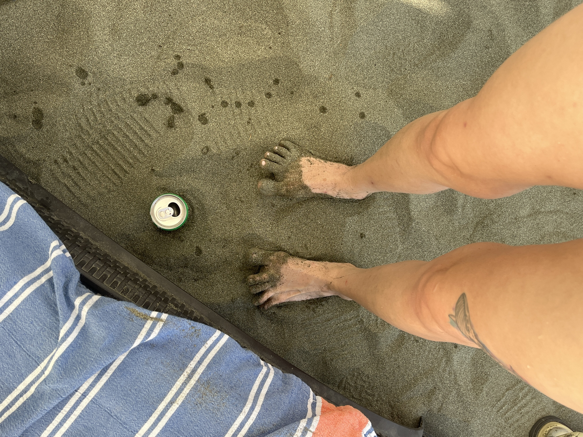 In Agios Tychona Community, Cyprus, a person's sandy feet are the central focus of this image, taken from a high-angle perspective. The sand is damp and speckled with darker wet spots, forming distinct toe impressions from the left foot and some smudges from the right. A partially open, green and silver soda can rests on the sand to the left of the feet. A blue and white striped towel with an orange stripe is draped on the left side of the frame, partially obscuring a textured black surface. The person's legs and knees are visible, with a tattoo of a stylized fish on the left thigh. The lighting suggests an outdoor, possibly sunny environment.