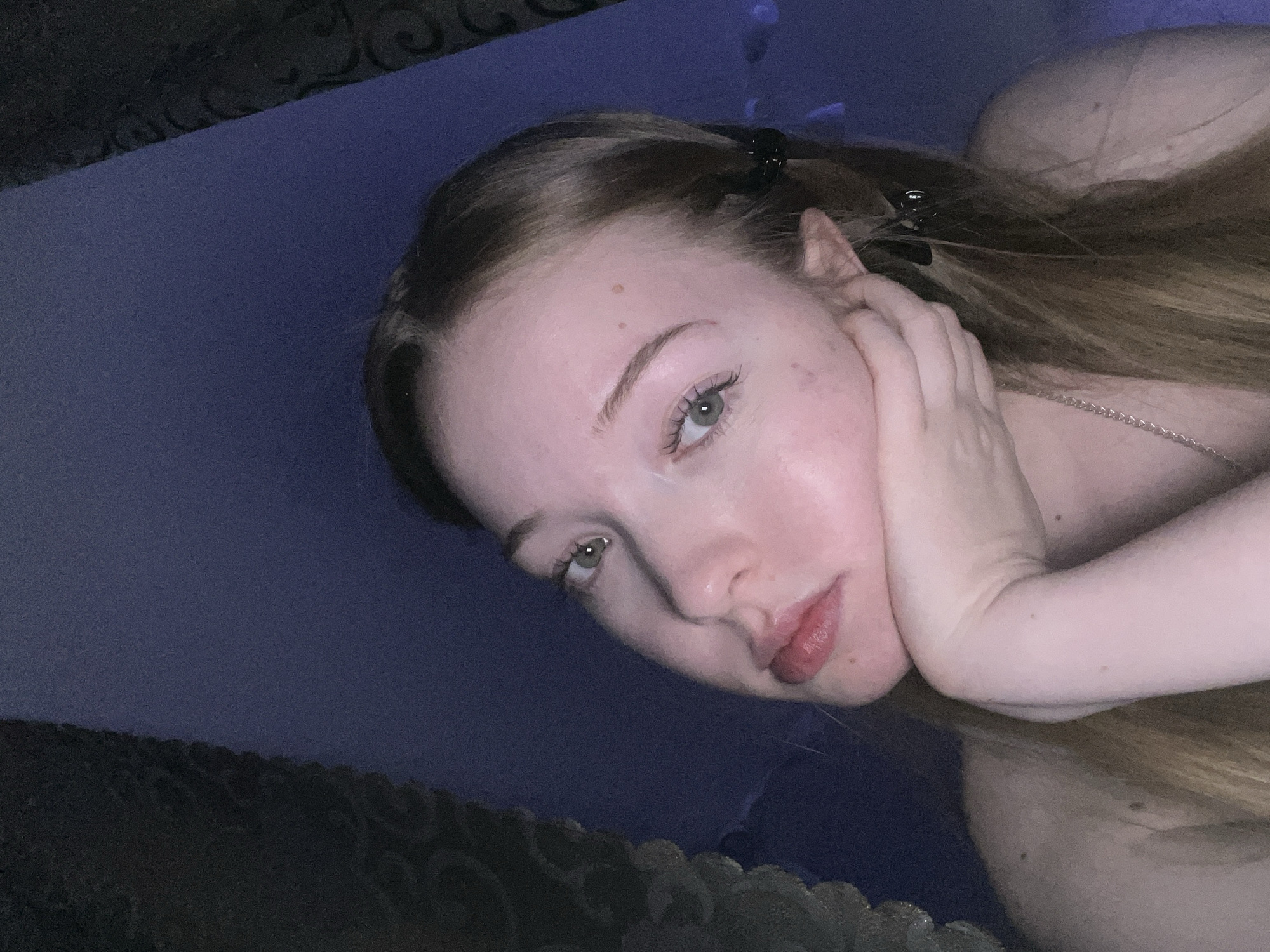 A close-up selfie shows a young, fair-skinned woman with light brown hair. Her hair is partially held back by a black clip. She rests her face on her hand, with a silver chain visible on her neck. The background appears to be a dark blue or purple surface, possibly a bed or couch. The image has low lighting. There is no visual evidence indicating the location to be the city of Chaikovsky, Russia.