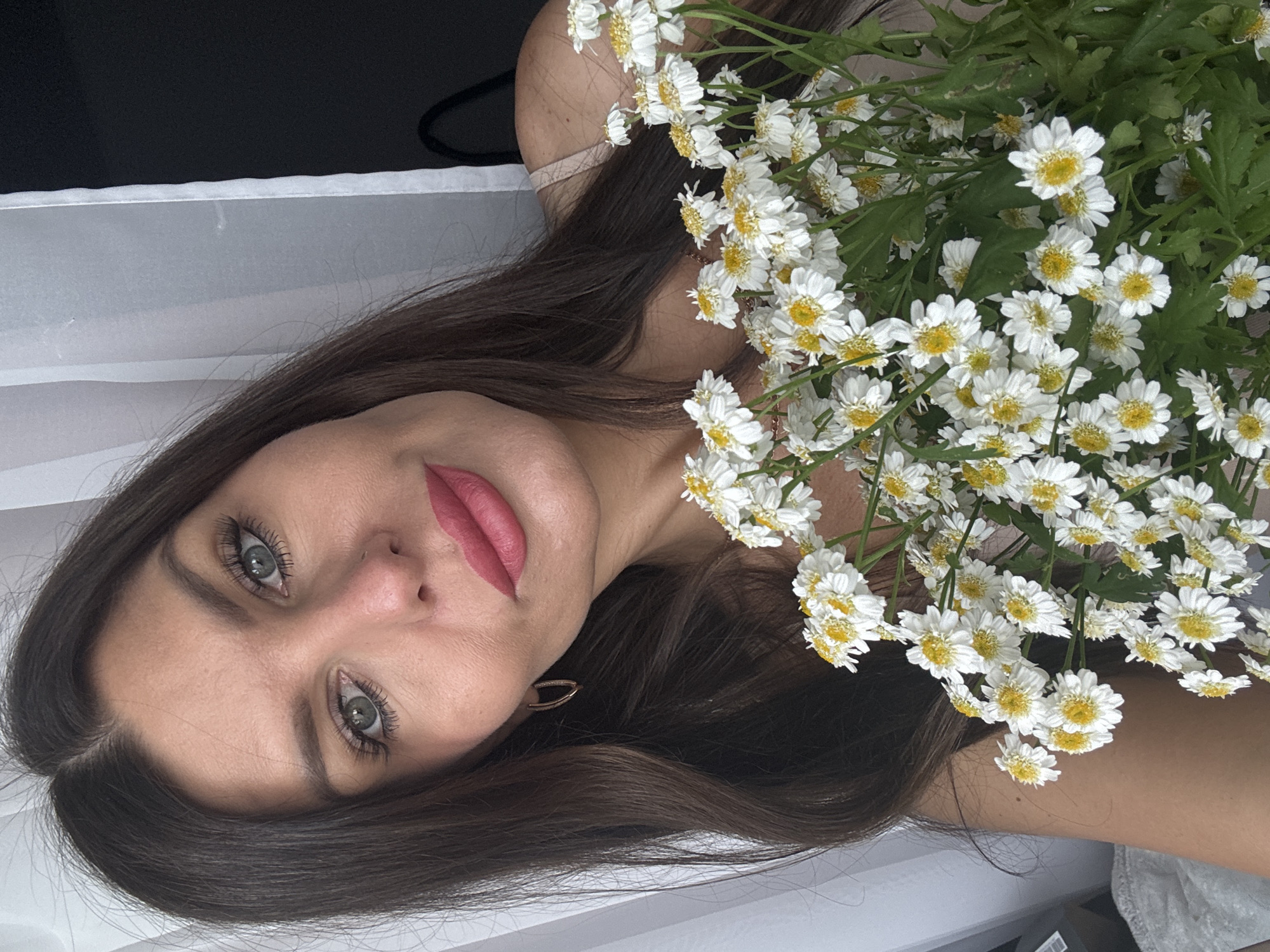 A medium shot shows a woman holding a bouquet of white daisies with yellow centers. She has long, dark brown hair, light green eyes, and is wearing pink lipstick. Her head is tilted slightly, and she is looking towards the camera. She is also wearing a pair of gold hoop earrings. The background consists of a white curtain on the left and a dark surface on the right. The image is taken indoors, with soft lighting. There is no discernible connection to the city of Chaikovsky, Russia.