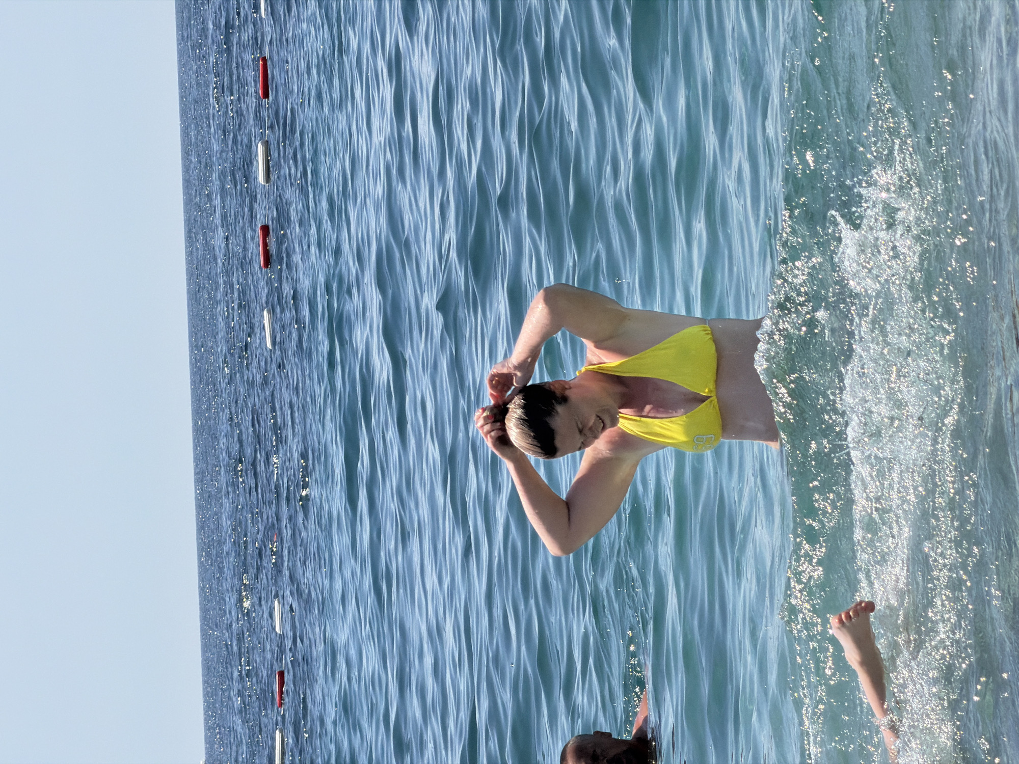 The scene captures a woman in a yellow swimsuit standing in the water in Boreti, Montenegro. The water has a clear blue color. A swimming lane line with alternating red and white floats is visible in the water to the left. The woman has her hands raised to her head, apparently fixing her hair. Another person is partially visible in the water toward the bottom of the image. Additionally, a pair of feet are seen closer to the surface of the water. Splashes surround the woman.
