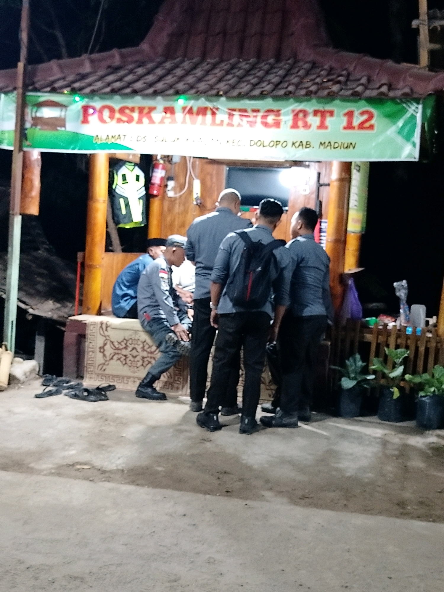 The image depicts a night scene outside a building with a tiled roof and bamboo pillars, likely a community post or local establishment. A banner above the entrance reads "POSKAMLING RT 12" and below it "ALAMAT: DS. SULUK KEC. DOLOPPO KAB. MADIUN". Several individuals are present. In the foreground, three men in dark shirts and pants stand facing away from the camera. One of them is wearing a backpack. To the left, another man in a grey shirt and dark pants is seated on a patterned mat, with several pairs of sandals on the ground nearby. A person in a blue shirt is also seated. A reflective vest is hanging on a pillar. The environment appears to be outdoors, with a dirt or concrete ground. Various items, including plants in pots, are visible to the right of the frame. The lighting suggests it is nighttime.
