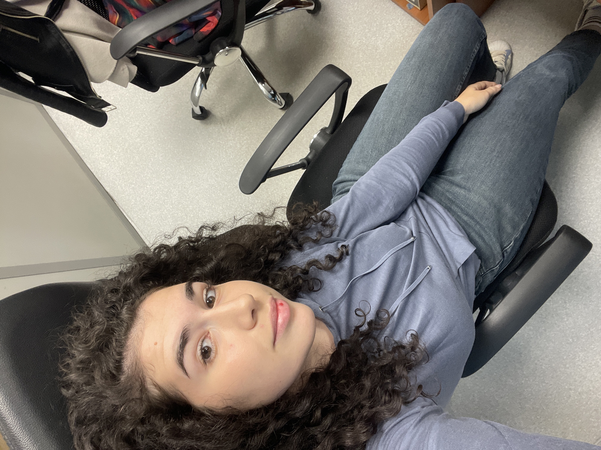 A young woman is depicted reclining in a black office chair, looking directly upwards at the camera. She has voluminous dark curly hair, light skin, and brown eyes, with a small red mark visible on her upper lip. She is dressed in a light greyish-blue hooded long-sleeved top and blue jeans, with light-colored athletic shoes visible on her feet. Her right hand rests casually on her lap.
The setting is an indoor room, likely an office or institutional space, featuring light grey speckled flooring and plain light-colored walls. The chair she occupies has black upholstered components and armrests. In the background, another black office chair with a chrome base and wheels is partially visible, with a black jacket, featuring a colorful inner lining, draped over its back. The woman's relaxed posture suggests a casual moment, possibly a selfie or a candid photo taken from above. The scene is situated in Sofia, Bulgaria. No text is visible within the image.