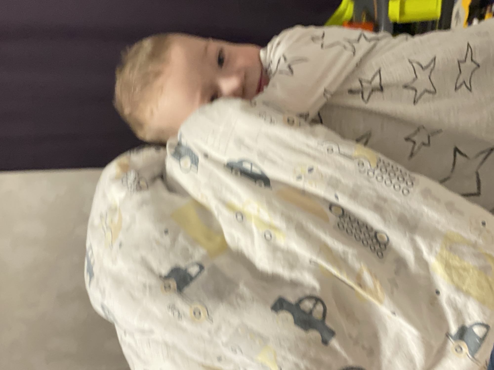 The image depicts a young child partially obscured by a patterned blanket in an indoor setting located in Sochi, Russia. The child, exhibiting light blonde hair and fair skin, has the right side of their face visible, including a blue eye and a portion of their cheek, looking towards the viewer with a neutral expression. The left side of the child's face and upper body are concealed by a white blanket or duvet, which is adorned with grey and yellow patterns, specifically depicting stylized vehicles (such as cars and possibly construction equipment) and outline stars. The background consists of a blurred, light grey wall on the upper left and a blurred, dark purple curtain on the right. Faintly visible on the far right, beneath the curtain, are indistinct, brightly colored objects in yellow, green, and orange hues, suggesting storage or toys.