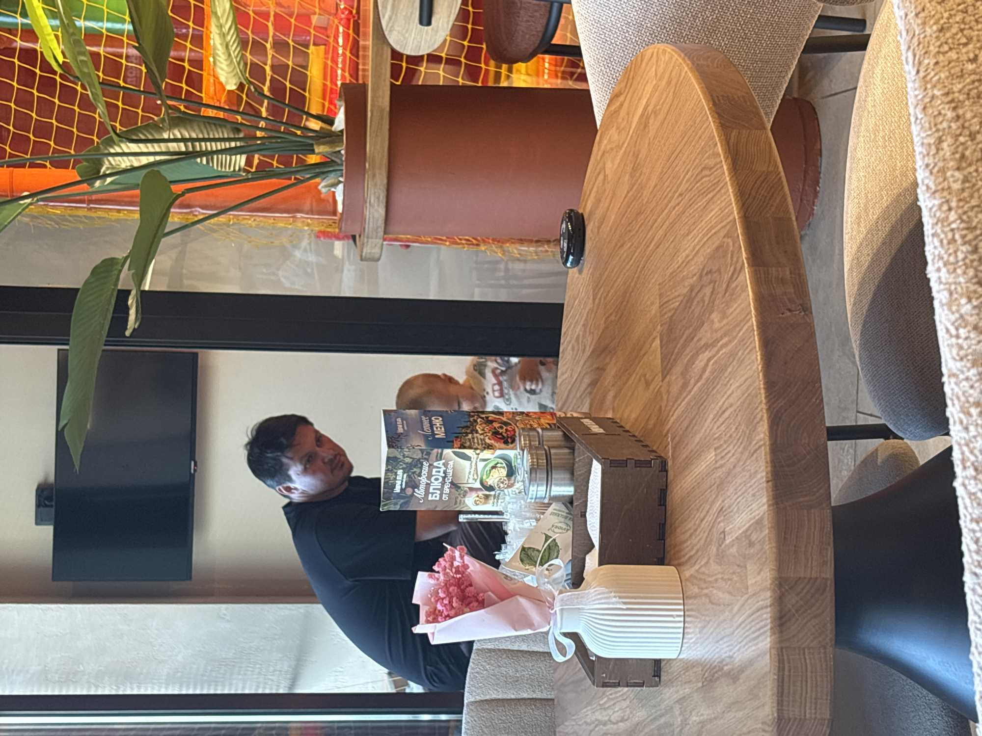 In an indoor dining establishment, a man in a black t-shirt is looking towards the camera while bending over. Beside him, a toddler wearing a patterned shirt is also looking down. On a wooden table in the foreground, a wooden tray holds a white vase-like object, a small bouquet of pink flowers, packets of condiments, and a menu. The man and toddler are positioned behind the table. The background features a large plant in a terra cotta pot, a television mounted on a wall, and a colorful play area with netting. The overall setting suggests a family-friendly restaurant. The scene appears to be located in Naberezhnye Chelny, Russia, given the presence of Russian text on the menu.