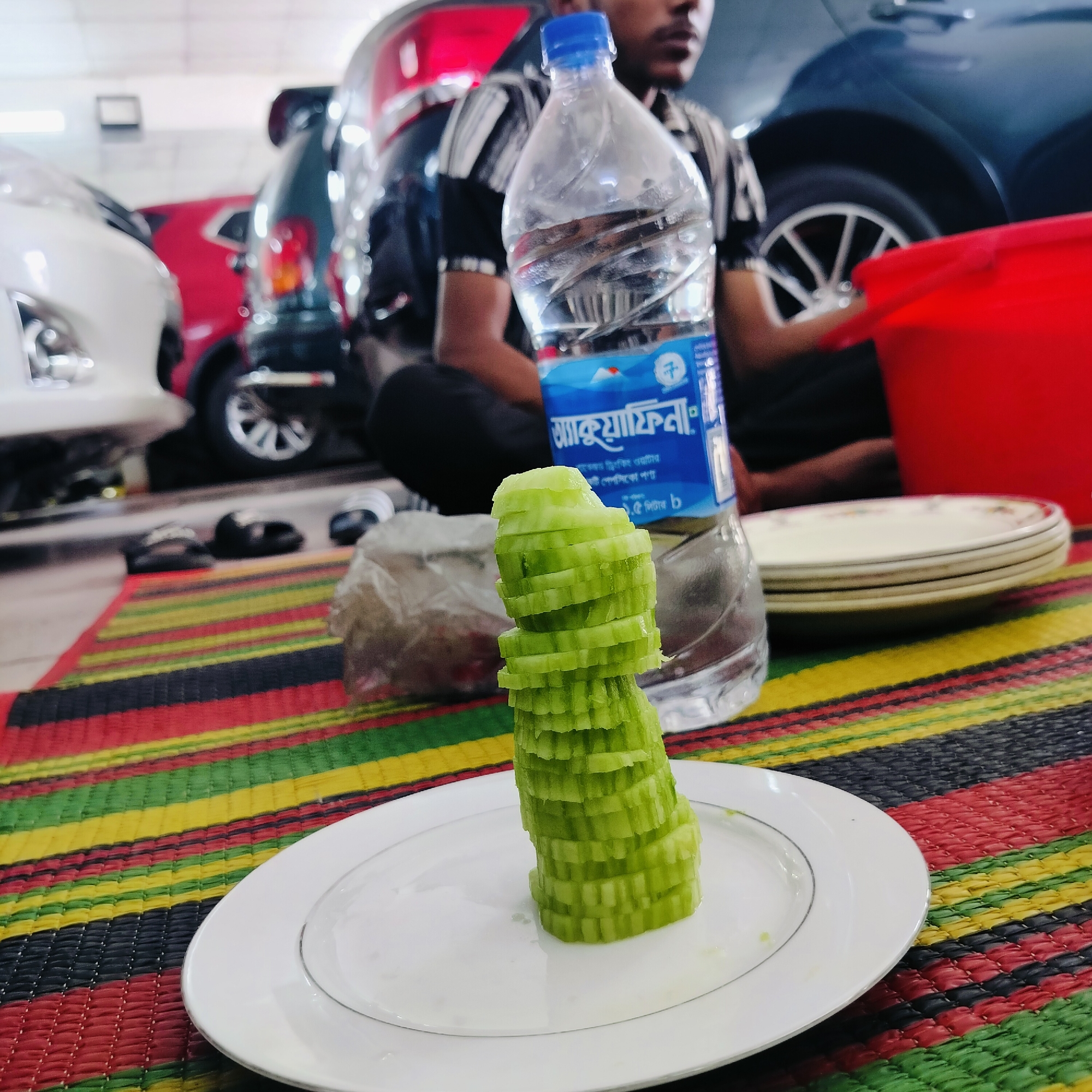 A man is sitting on a striped mat in what appears to be a garage in Dhaka, Bangladesh. In front of him is a plate with sliced cucumbers stacked into a tower, a bottle of water, and a stack of plates. There are several cars parked in the background. The general environment is indoors, possibly a car repair shop or garage due to the presence of multiple vehicles.