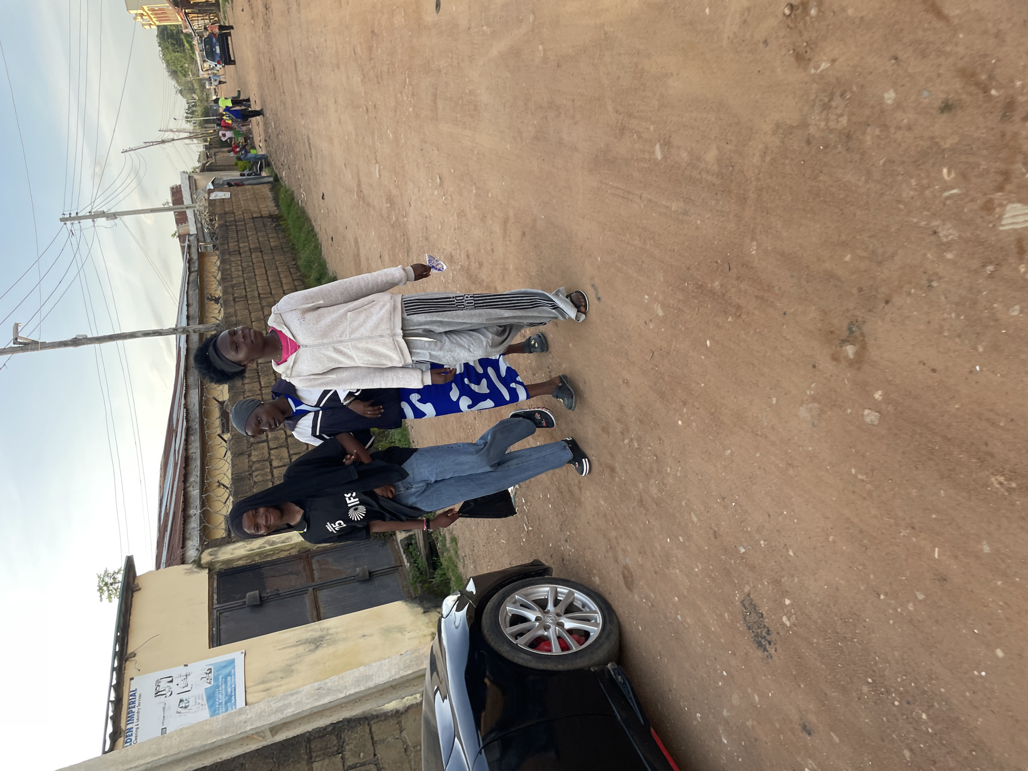 The image depicts a street scene in Barakin Akawo, Nigeria. Three young women are walking on a dirt road. The woman on the left is wearing a black t-shirt with white lettering and a black headscarf. She has on light blue jeans and black slip-on shoes, and is carrying a black plastic bag in her right hand. The middle woman is wearing a blue and white patterned dress and grey slip-on sandals. The woman on the right is wearing a light gray hooded sweatshirt and gray track pants with black stripes. She is barefoot. To the left of the women is a black car. The background shows a building with a sign that says "GOLDEN IMPERIAL Cleaning & Laundry Services," a brick wall, and power lines. Further down the street, other people and vehicles are visible. The ground is composed of reddish-brown dirt and scattered debris. The sky is overcast with a soft light, suggesting it is late afternoon or early morning.