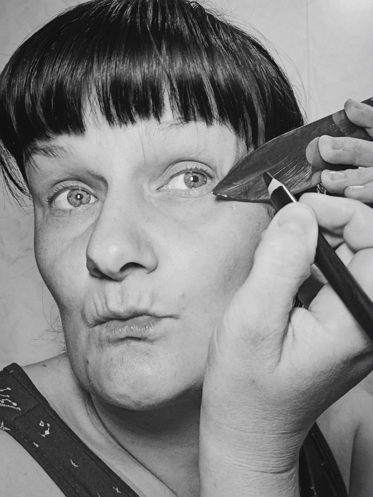 A black and white close-up image depicts a person applying eyeliner. The person has short, dark hair with bangs and is making a pouting expression. In the right hand, they hold a black eyeliner pencil and appear to be applying it to the right eye. In the left hand, they hold a flat, rectangular object, possibly a makeup tool or straight razor, near the eye to guide the eyeliner application. The person is wearing a tank top with a zodiac sign printed on it. The background is a light, neutral color. The location is Unknown, Unknown.