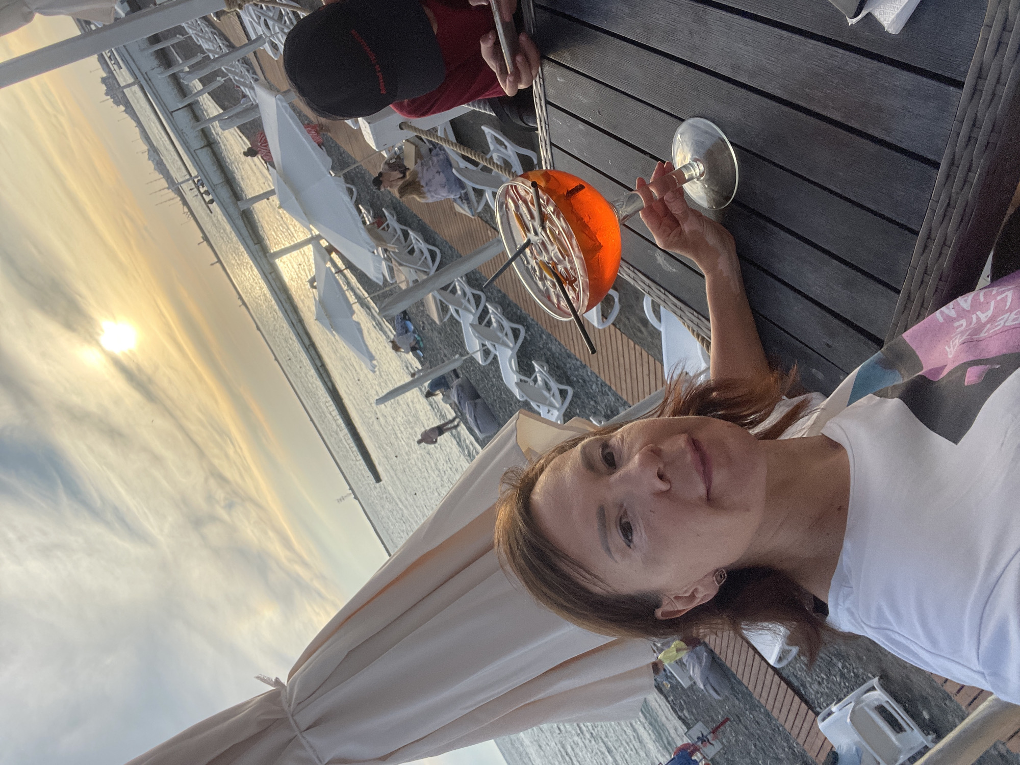 A woman with medium-length brown hair is taking a selfie at a beachside cafe in Sochi, Russia. She is wearing a white t-shirt. In front of her on a dark wooden table is a large glass of orange aperitif with two straws and citrus slices. To her right, a person wearing a red shirt and a black cap is using a smartphone. In the background, lounge chairs, umbrellas, and people are visible on a pebble beach overlooking the Black Sea. The sun is setting, casting a warm glow on the scene.