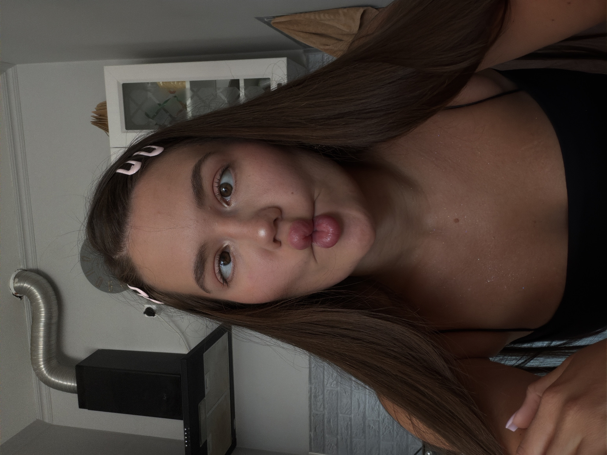 The image shows a close-up, rotated portrait of a young woman with long, dark brown hair. She is wearing a black top, and her right shoulder and chest are visible, dusted with glitter. The woman is making a pouty face with her lips pursed. Two light pink butterfly clips are visible in her hair near her forehead.
In the background, on the left, a stainless steel ventilation pipe curves upwards. Below it, a black kitchen hood is mounted on a white wall. To the right of the hood, a portion of what appears to be kitchen cabinetry with a white frame and glass inserts is visible. The lighting suggests it is daytime, but the indoor setting makes it difficult to determine the exact time. The overall scene appears to be an indoor domestic environment, possibly a kitchen. The woman's expression is playful and somewhat cheeky. There is no visible text in the image.