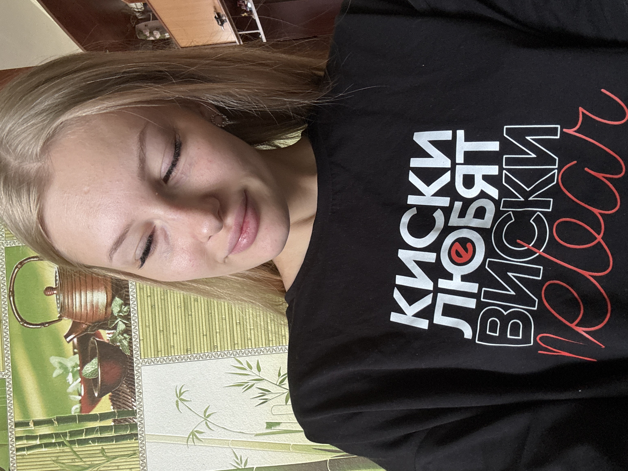 A young blonde woman with light skin and her eyes closed is positioned in the center of the frame, facing slightly to her right. She is wearing a black crewneck sweatshirt with white text that reads "КИСКИ ЛЮБЯТ ВИСКИ" and red script text underneath that appears to say "relax". She has on small, hoop-like earrings. The background consists of a wall with a repeating bamboo and tea set pattern. The lighting suggests an indoor setting, possibly a home. There is no indication of the city of Комарихинский, Russia.