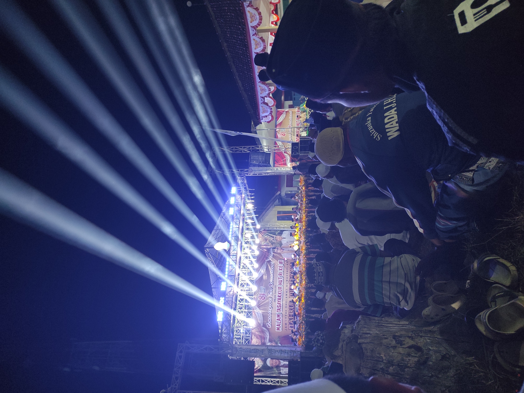 An outdoor nighttime event is depicted, with a stage illuminated by numerous spotlights projecting beams of light into the dark sky. A large banner on the stage reads "MALAM TASAYAKKUR HUT RI KE 81" and other Indonesian text. Several men are seated on the stage, some appearing to be speaking or leading the event. In front of the stage, a crowd of people, predominantly men wearing Muslim attire such as peci and sarongs, are seated on the ground. Some individuals in the foreground are facing away from the camera, while others are partially visible from the back. Shoes and sandals are scattered on the ground in the lower portion of the image. The environment appears to be a grassy or dirt field.