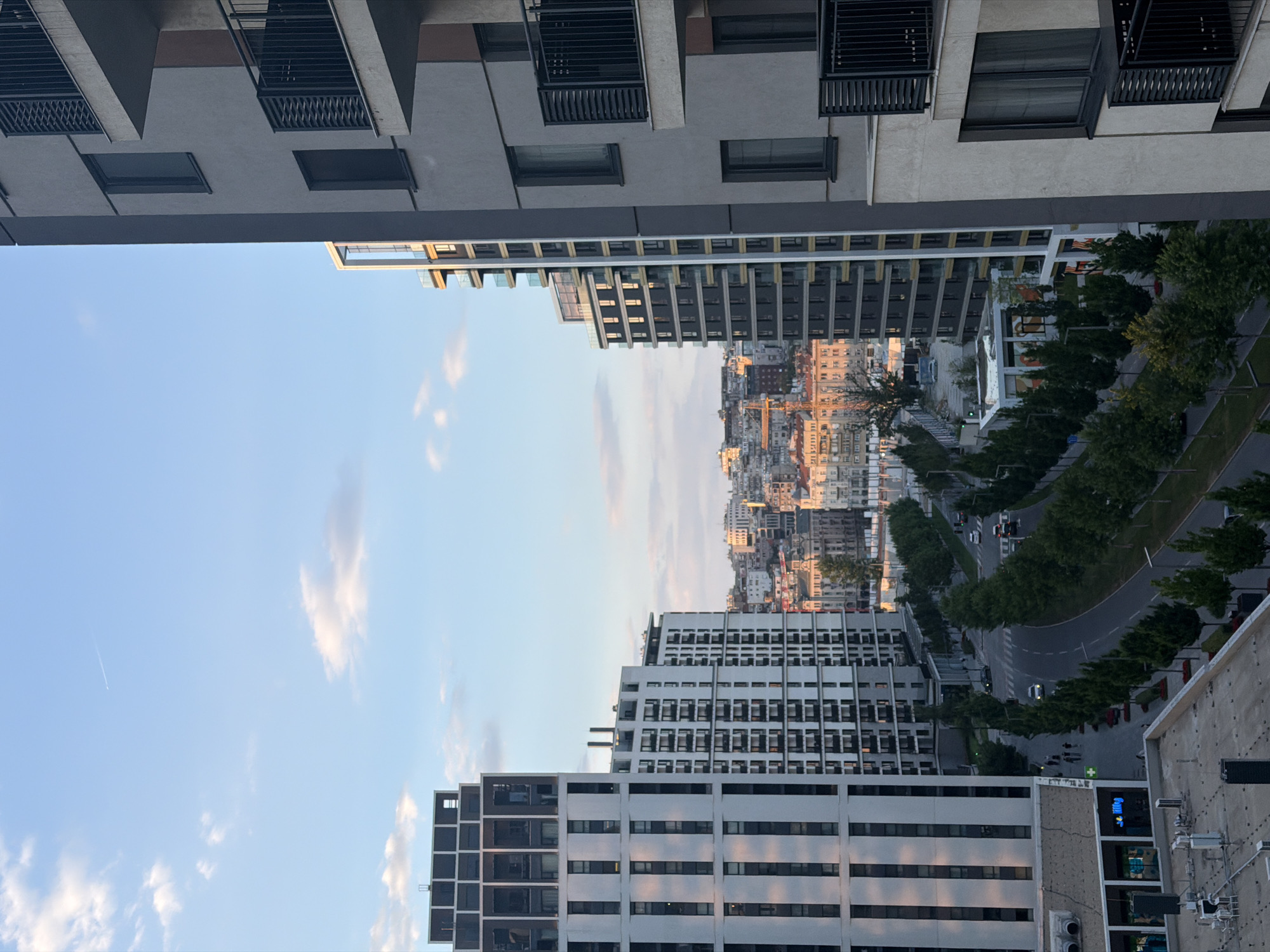 The image depicts a modern urban landscape in Belgrade, Serbia, with a focus on contemporary architecture. In the foreground, two tall residential buildings dominate the scene. The building on the left is a white high-rise with a distinct grid pattern of dark windows and vertical accents. The building on the right is a grey structure featuring multiple balconies with black railings.
Between these buildings, a tree-lined street curves through the cityscape. Several cars are visible on the road, suggesting moderate traffic. Pedestrians are also present, walking along the sidewalks.
In the background, a dense collection of older, traditional buildings with varied architectural styles and colors forms the city's skyline. A prominent construction crane can be seen among these older structures, indicating ongoing development. The sky above is a pale blue, with scattered white clouds and a faint contrail from an airplane. The lighting suggests it is late afternoon or early evening, with the sun casting warm tones on some of the distant buildings. The overall impression is one of a city blending new and old urban development.