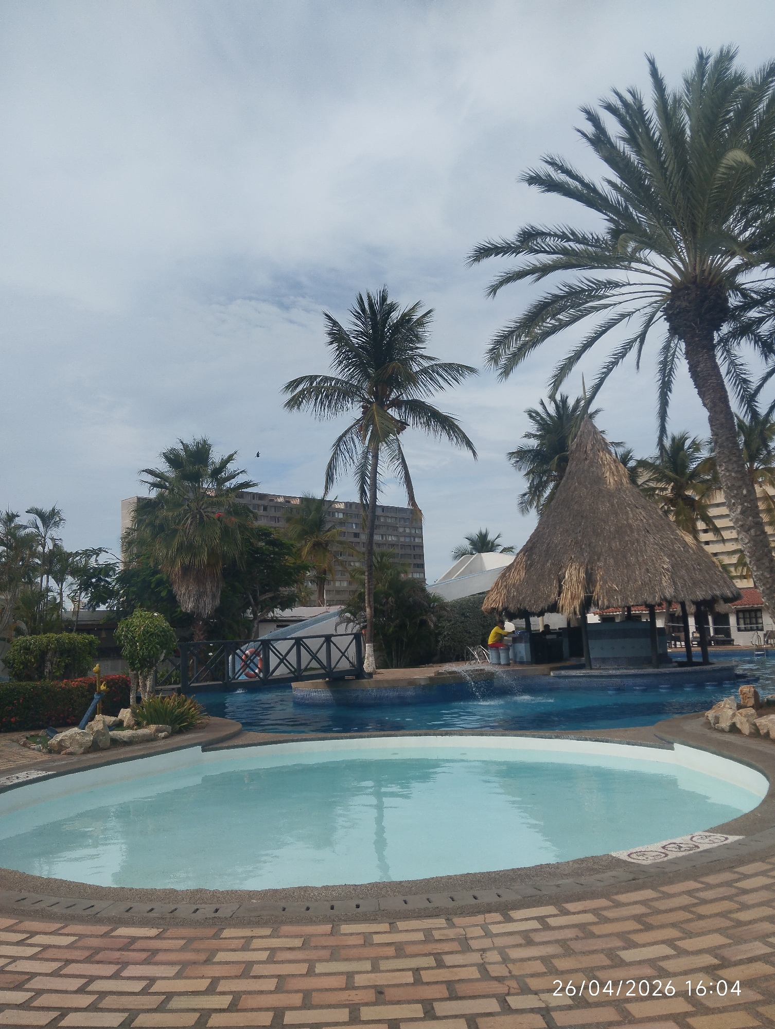 A daytime outdoor shot shows a resort swimming pool with a shallow circular pool in the foreground, a larger pool with a thatched-roof bar, palm trees, and a multi-story building in the background. The foreground pool is filled with light blue water, surrounded by a brick-paved area. Beyond the pool, there is a larger swimming pool with a small bridge and a thatched-roof bar with a person in yellow serving. Palm trees are scattered throughout the scene. In the background, a tall, light-colored building is visible under a cloudy sky. The location is in the city of Unknown, Unknown. A date stamp of "26/04/2026 16:04" appears in the lower right corner.