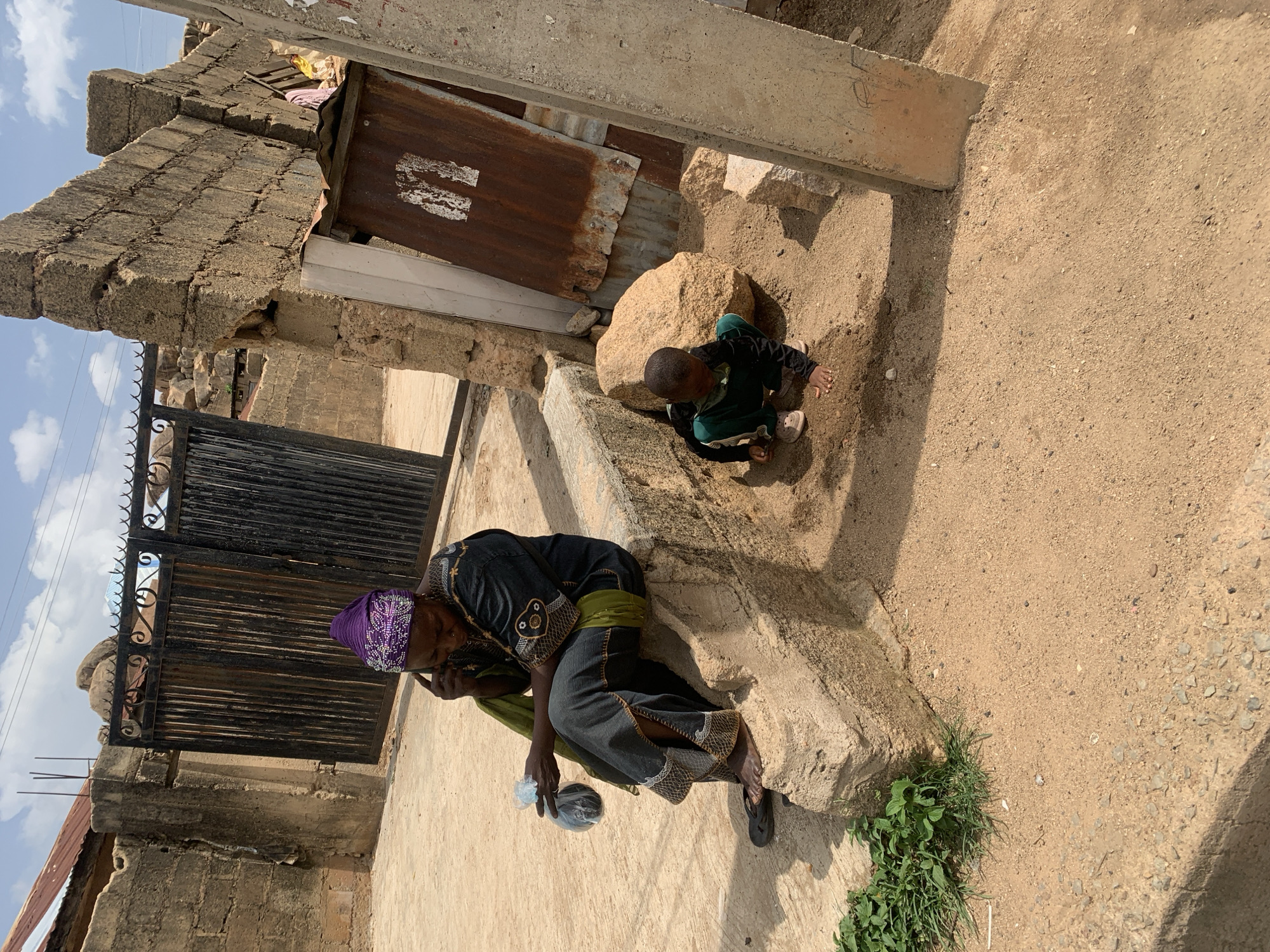 Outside on a sandy lot in Jos, Nigeria, an older woman wearing a dark patterned dress and purple head covering sits on a concrete step. She holds a plastic bag. A young child in a dark outfit plays in the sand near a large rock. The area is surrounded by buildings, some with rusted metal siding and stone walls. A black metal gate is visible in the background. The sky is partially cloudy.
