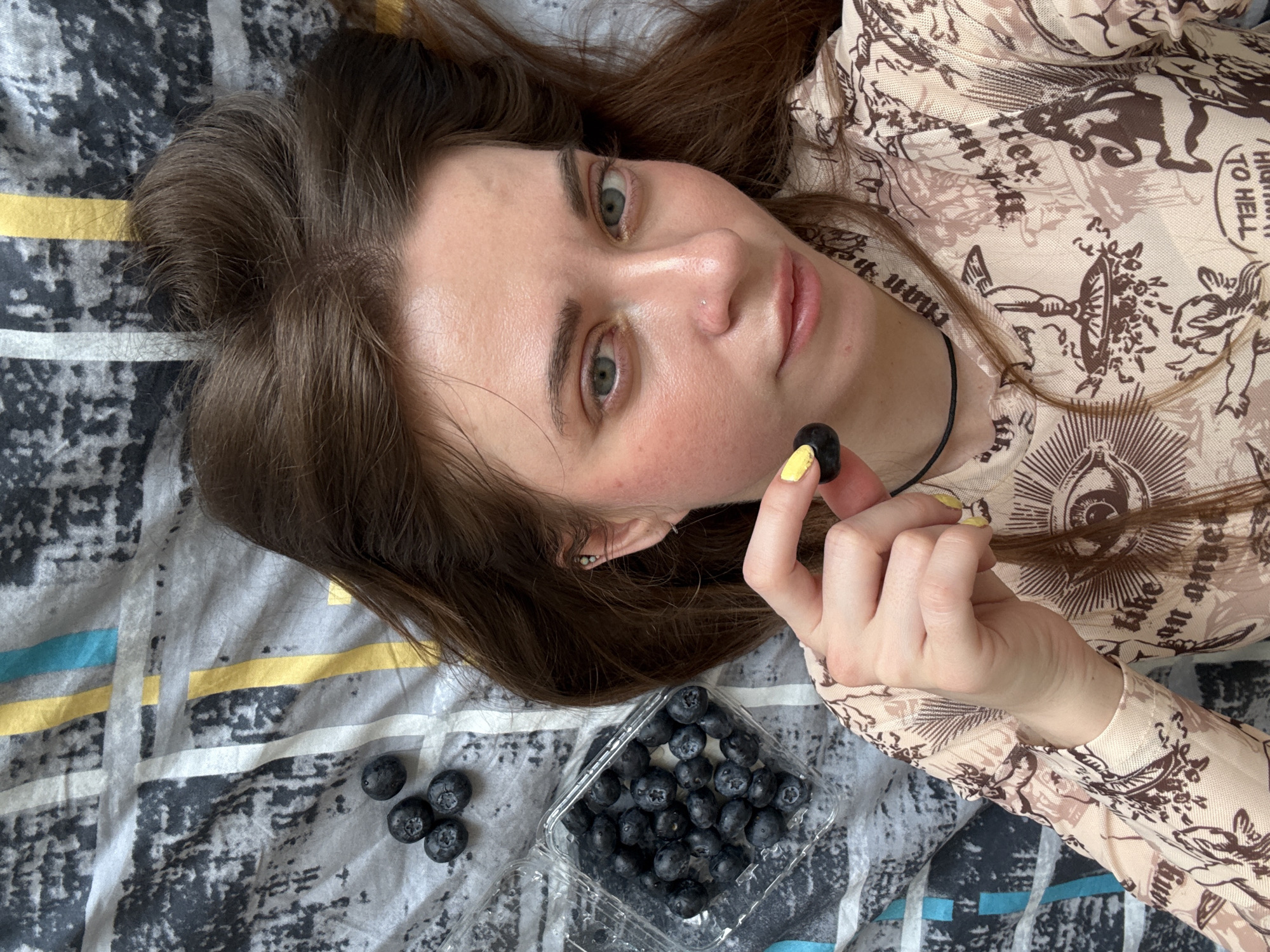 A young, fair-skinned woman with long brown hair is lying on her back on a patterned bedspread. She is holding a single blueberry in her right hand, and a plastic container filled with blueberries is next to her on the bed. Several loose blueberries are scattered near the container. She is wearing a long-sleeved shirt with a light beige print that includes text and illustrations. The scene does not appear to be located in Moscow, Russia; there are no visual clues to support this.