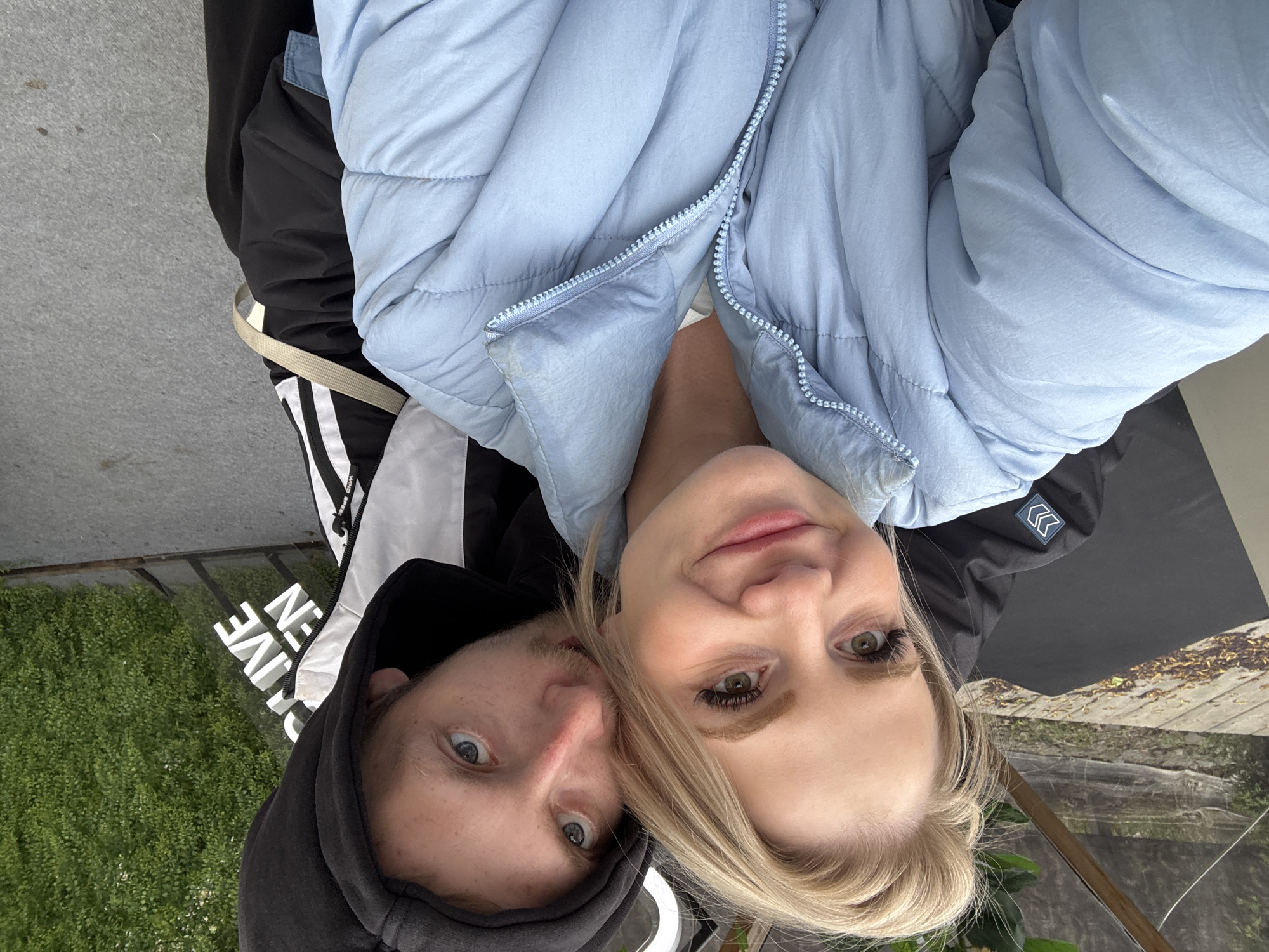 A selfie captures a couple, a man and a woman, close together with their faces prominent in the frame. The woman has blonde hair and is wearing a light blue puffer jacket, partially unzipped. Her facial expression is pleasant, with a slight smile. The man, positioned next to her, is wearing a black hoodie. Their faces are close, almost touching.
Behind them, the background reveals a glimpse of what appears to be a city scene. There is a reflection of greenery, possibly trees or bushes. Also, there is a signage that is blurred, but seems to have letters.
Without more information, the specific location in Saint Petersburg, Russia is not identifiable.