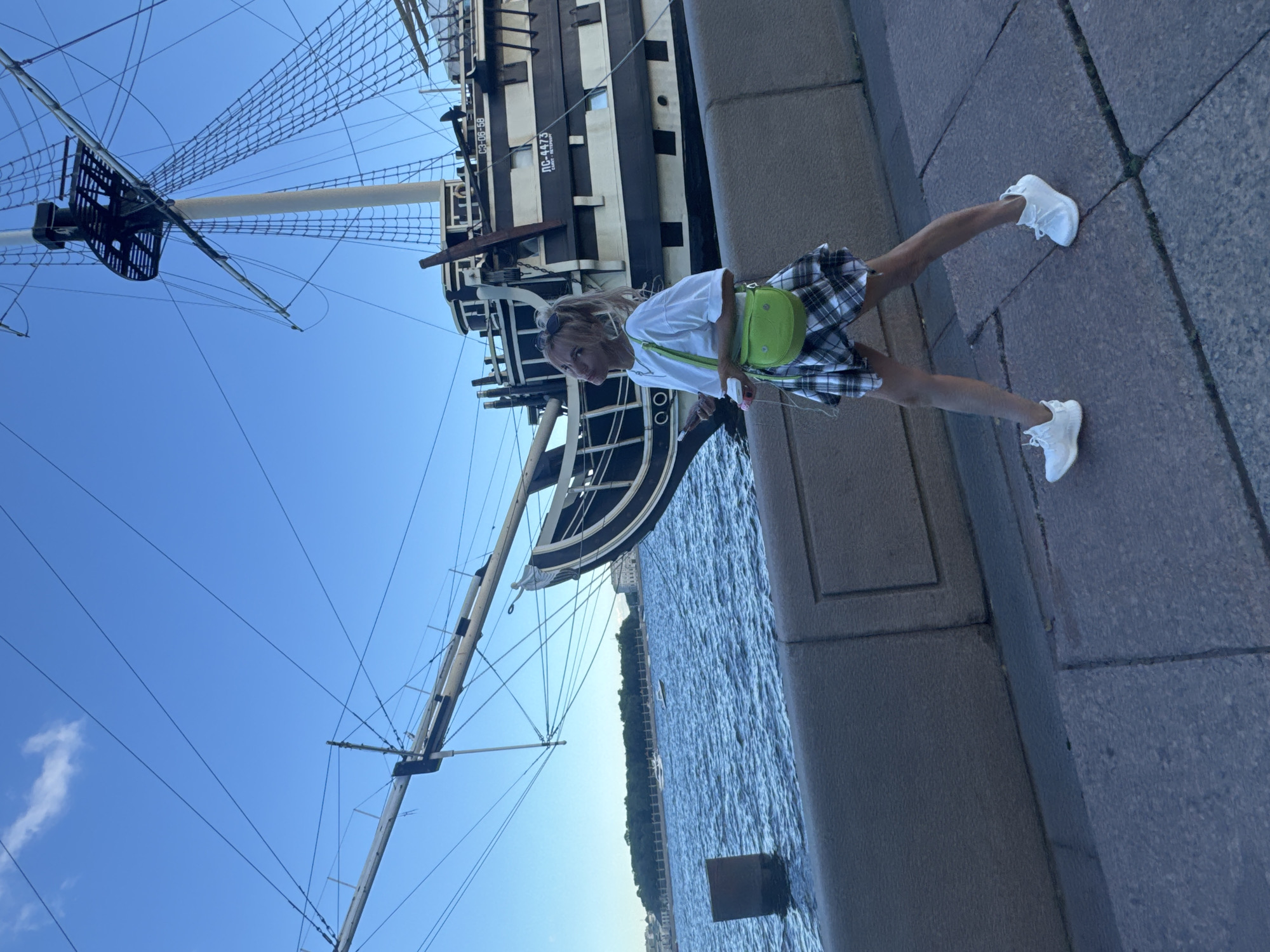 A young blonde woman wearing a white t-shirt, a plaid skirt, and white sneakers walks along a stone embankment next to a large wooden ship in Saint Petersburg, Russia. She has a bright green crossbody bag slung over her shoulder and is holding a phone in her left hand. The ship, which appears to be a historical replica or museum piece, is docked on the Neva River. The sky is clear blue with a few wispy clouds. The embankment is paved with large stone slabs, and a gray concrete wall borders the river. In the background, across the river, is a treed area with buildings. The image appears to have been taken in the late afternoon or early evening, as the light is soft and the shadows are long.