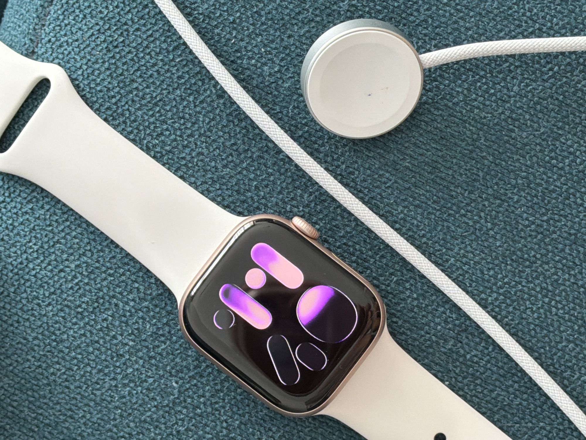 An Apple Watch and its charger are laid out on a teal textured surface. The watch face displays a vibrant, abstract graphic with purple and pink hues. The watch band is light-colored, likely white or cream, and appears to be made of silicone. The charger is a circular, silver magnetic puck with a white braided cable. The image is a close-up, top-down shot, with no discernible background elements to indicate a specific location within the city of Finestrat, Spain, or any other urban setting. The lighting is bright, suggesting it was taken indoors or in a shaded outdoor area. There are no people or animals present in the scene.