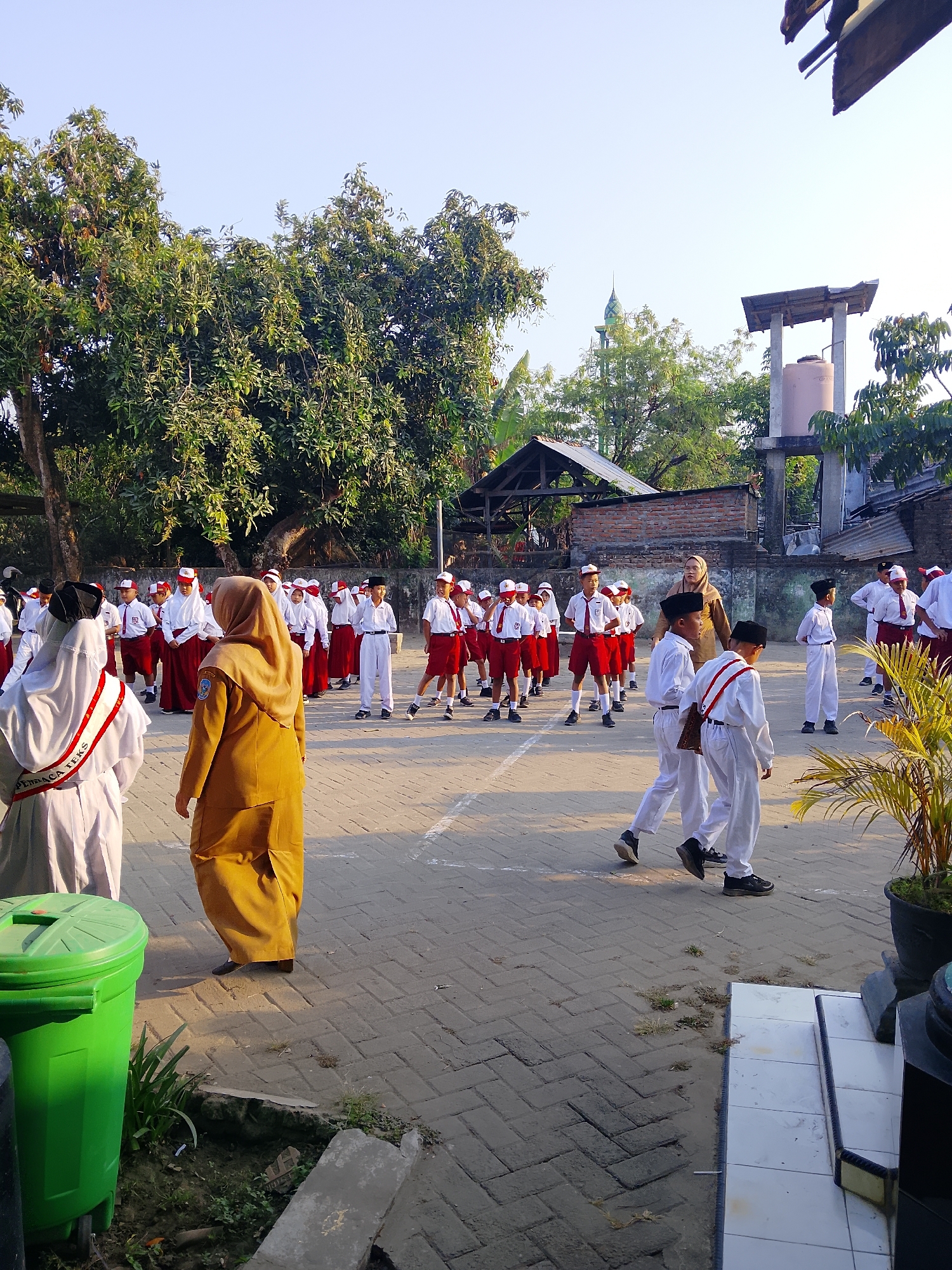 The image depicts an outdoor scene at what appears to be a school in Kepuh, Indonesia. A group of children in school uniforms, consisting of white shirts, red skirts or shorts, and red and white hats, are lined up and possibly participating in an activity. Some of the children are wearing black headwear. Two adults are also visible; one is wearing a tan-colored hijab and a long tan dress, and the other is wearing a white hijab and a white robe with a red sash. The ground is paved with interlocking bricks, and there is a large green trash bin on the left side of the image. In the background, trees, a building with a tiled roof, and a water tank on stilts are visible, along with what appears to be a minaret of a mosque. The lighting suggests it is daytime, likely morning due to the angle of the sun and the clear sky.